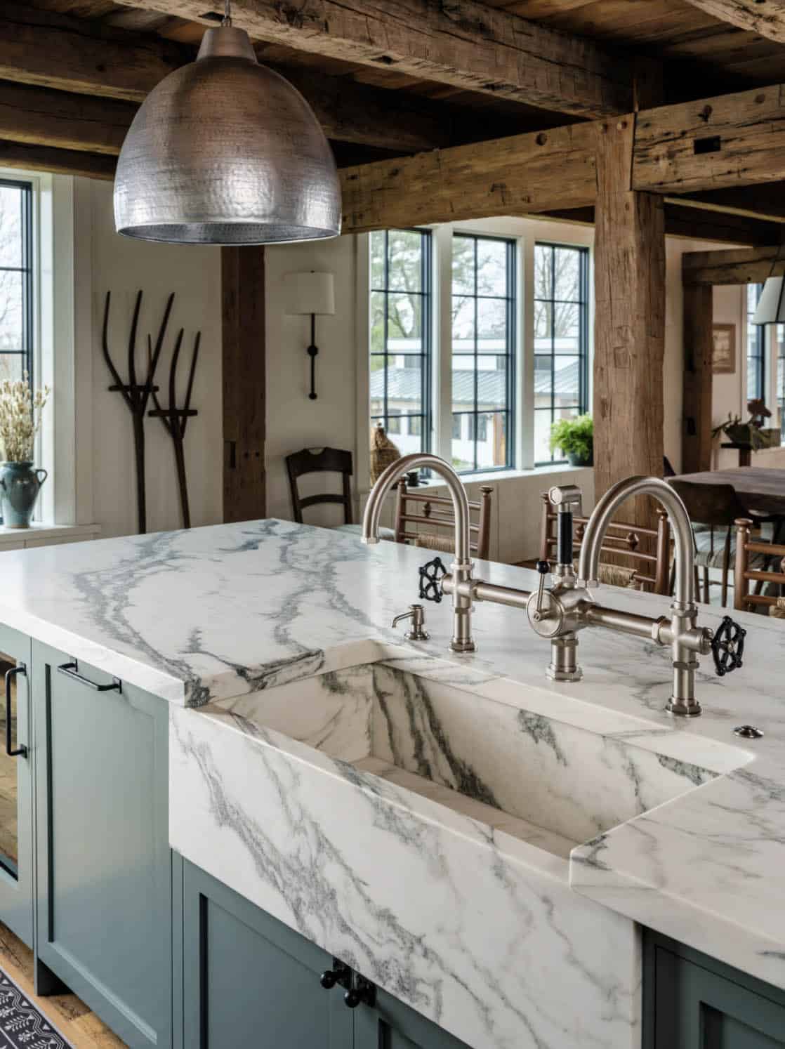 Close-up of barn kitchen island with dramatic book-matched marble waterfall countertop and apron-front farm sink, dual bridge faucets with industrial wheel handles, teal cabinet base, and hammered pendant above