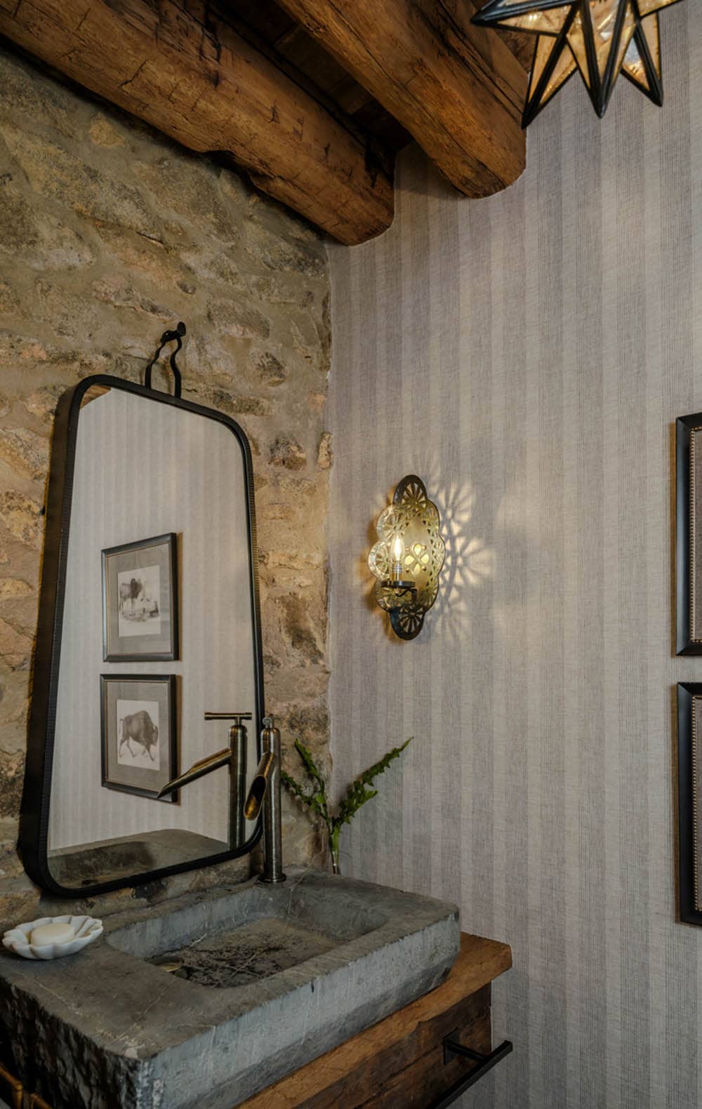 Powder room with carved stone trough sink on a wood vanity shelf, antique bronze waterfall faucet, iron-framed mirror, ornate brass wall sconce casting decorative shadow pattern, original fieldstone wall, and striped wallpaper