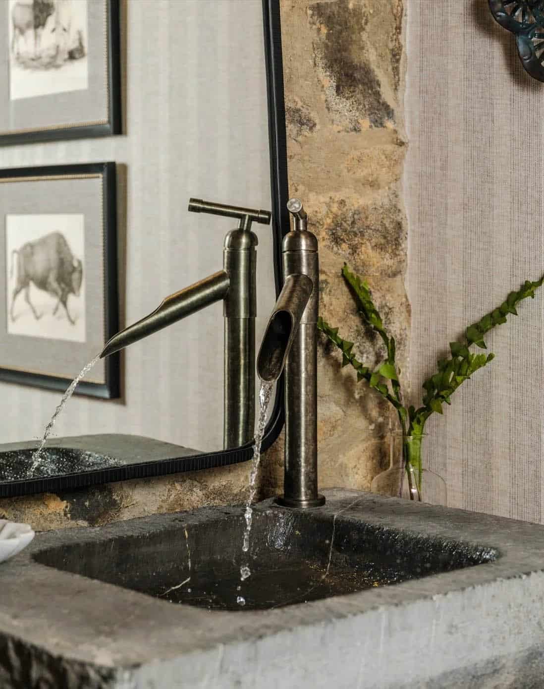 Close-up of powder room antique bronze waterfall faucet running water into a rough-hewn carved stone trough sink, with original fieldstone wall and striped wallpaper in background