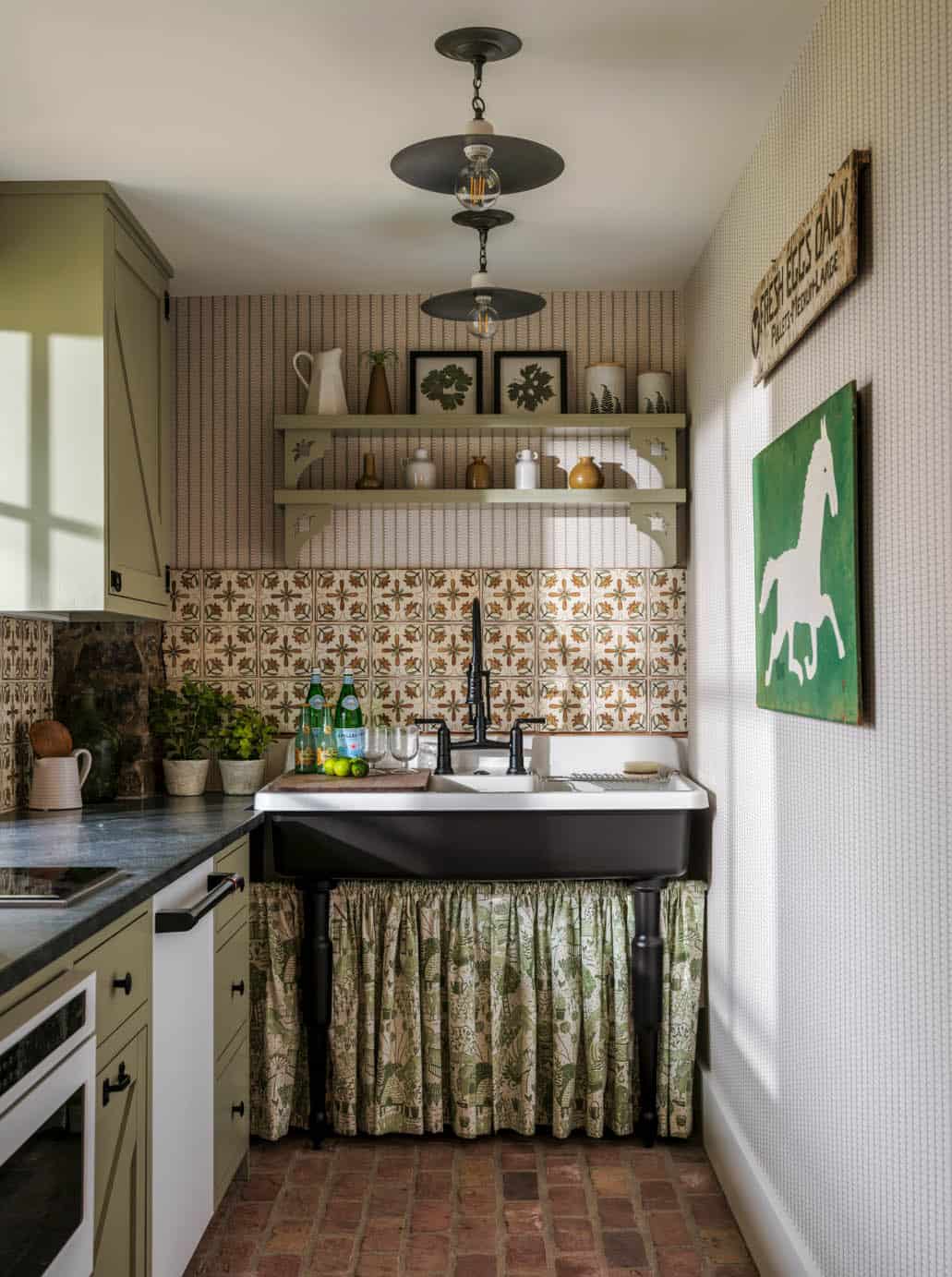 Kitchenette with sage green cabinetry, black apron-front farm sink with gathered floral skirt, decorative tile backsplash, painted open shelves, industrial globe pendants, terracotta brick floor, and horse folk art painting