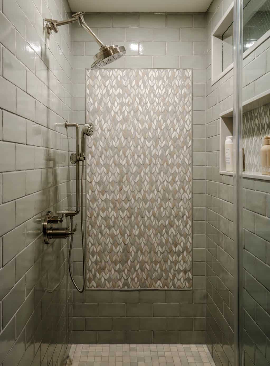 Walk-in shower interior with sage green subway tile walls, leaf-pattern mosaic accent panel, polished nickel rain head and hand shower on slide bar, built-in niche shelving, and small mosaic floor tile