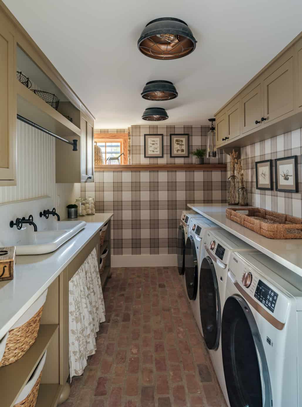 Barn home laundry room with plaid wallpaper, sage green shaker cabinetry, apron-front utility sink with black wall-mount faucet, gathered floral under-sink curtain, side-by-side washer and dryer, antique cage flush-mount lights, and terracotta brick floor