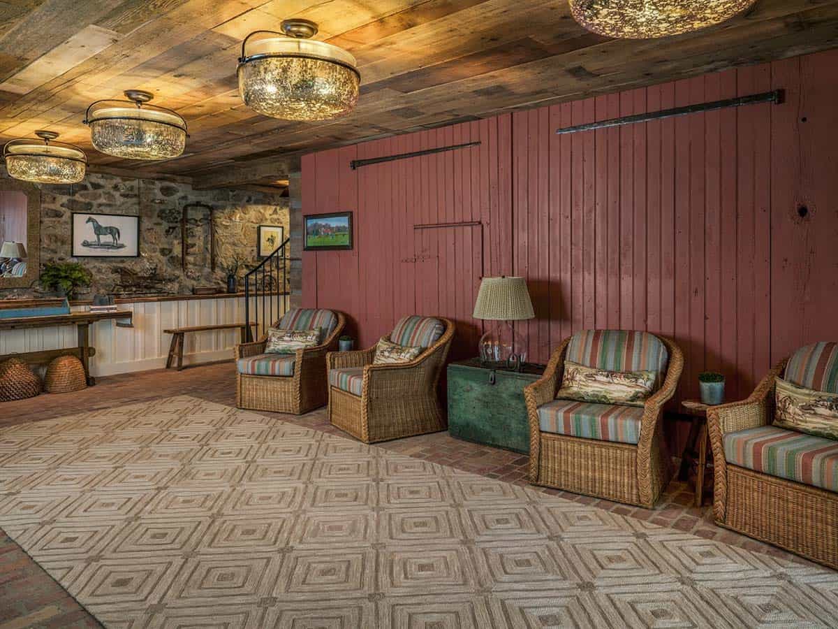 bank barn sitting room with original red-painted vertical board wall with sliding barn door hardware, wicker armchairs with striped cushions around a vintage painted trunk coffee table, geometric braided area rug, mercury glass flush-mount ceiling lights, reclaimed plank ceiling, original fieldstone wall, and framed horse prints