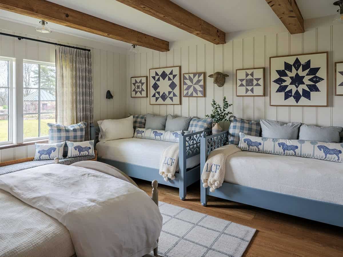 Children's bunk room with two blue-painted daybeds with trundle pulls, blue gingham and horse-print pillows, monogrammed throws, five framed barn quilt pattern prints on white beadboard wall, and original reclaimed timber ceiling beams