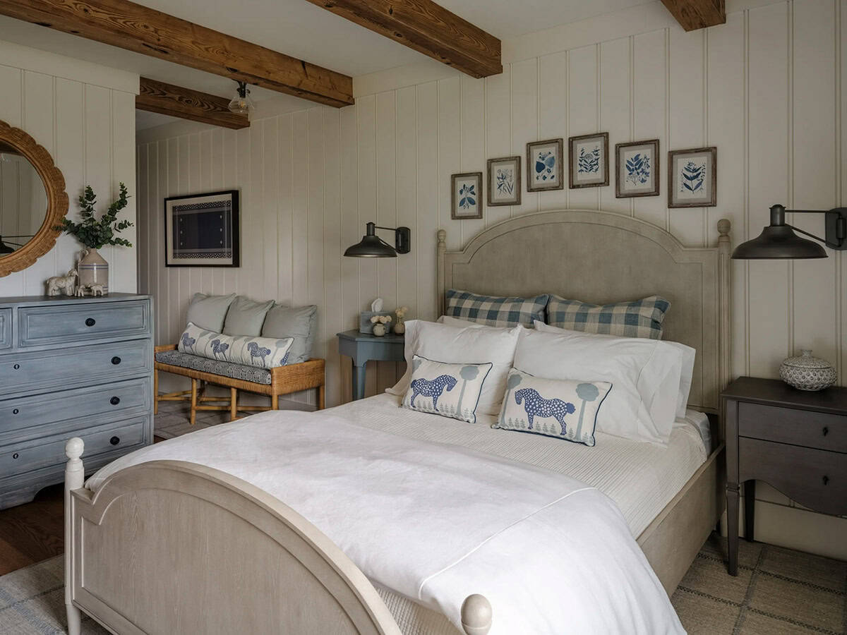 Guest bedroom in barn conversion with arched gray wood bed, blue gingham and horse-print accent pillows, blue-gray painted dresser, rattan bench, mismatched nightstands, black swing-arm sconces, row of framed botanical prints, and white beadboard walls with reclaimed timber beams