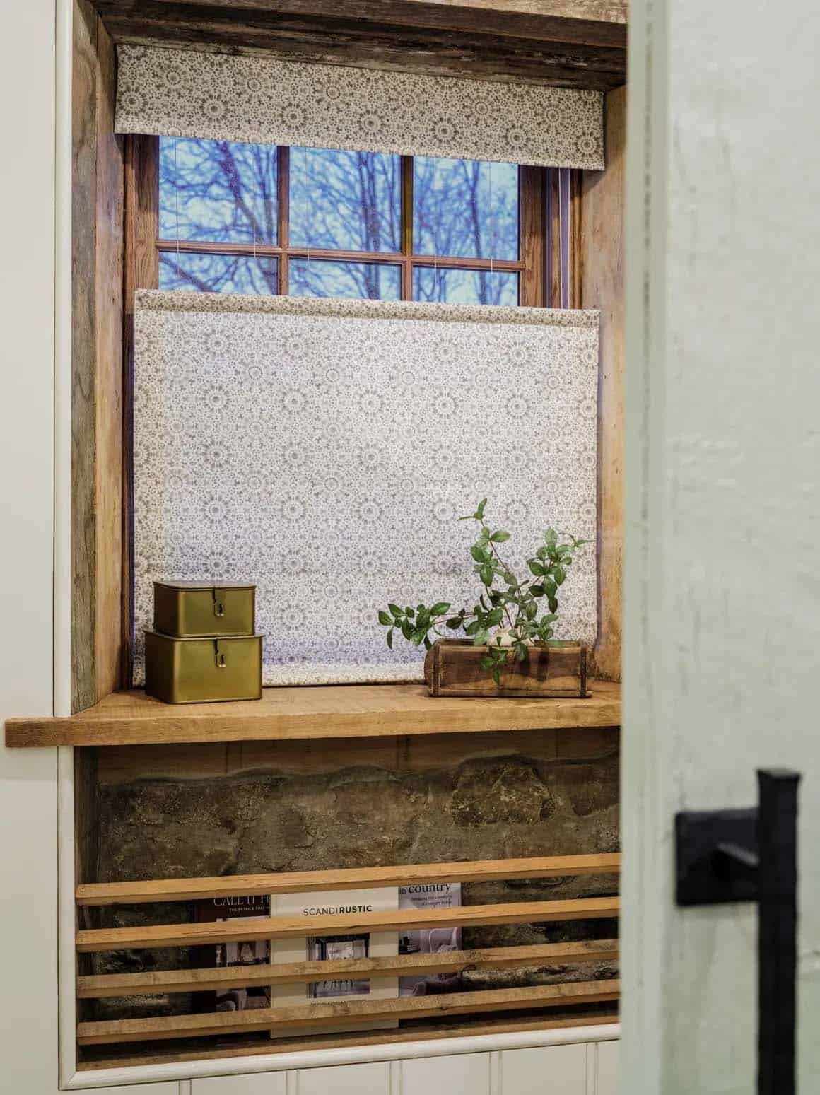 Deep-set original fieldstone window niche framed in reclaimed timber with a botanical-print Roman shade, wood shelf displaying brass tins and a greenery sprig, lower slatted wood magazine rack with design books, and black iron door hardware in foreground
