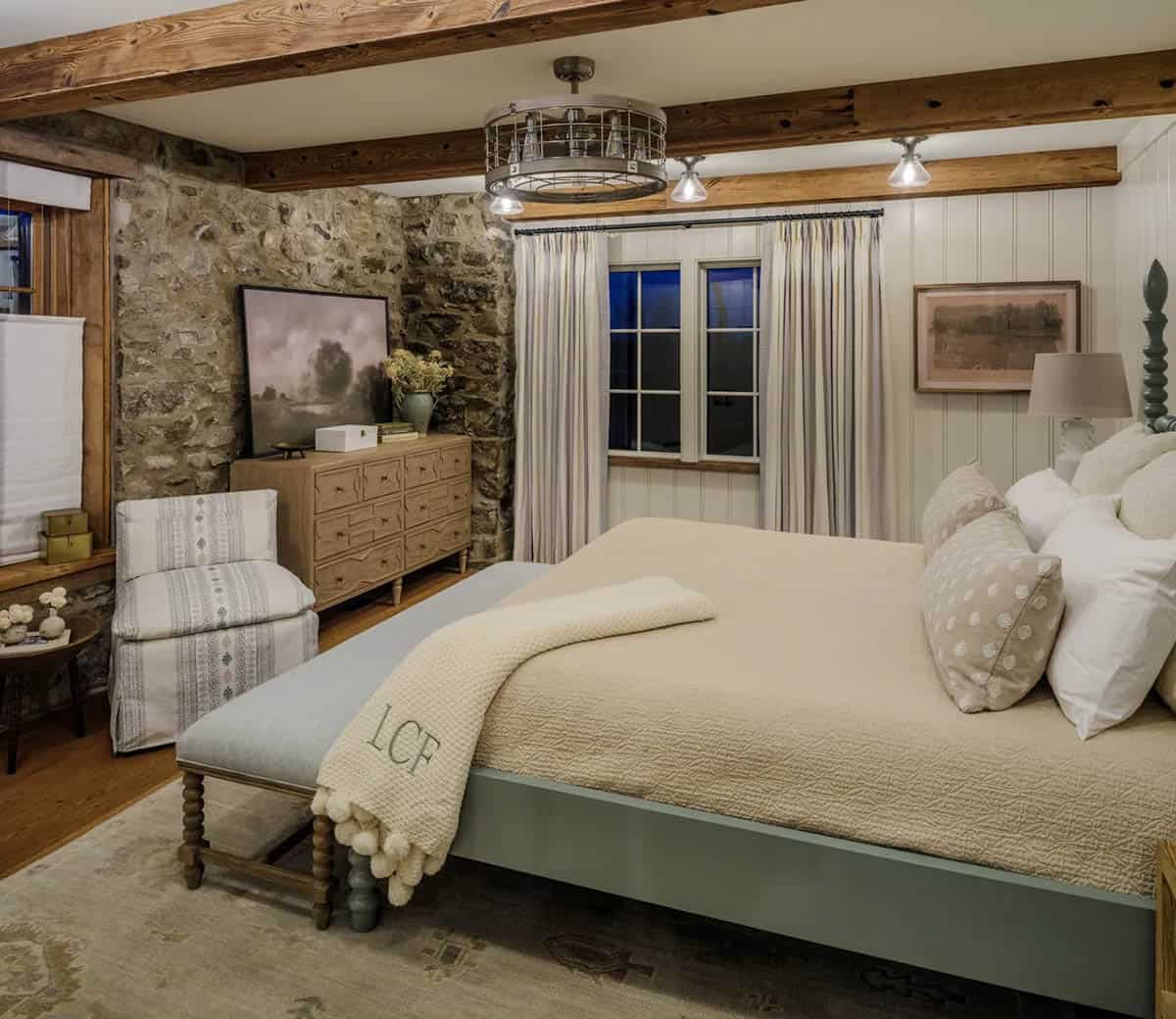 guest bedroom at night with sage green spindle bed frame, monogrammed cream throw, upholstered slipper chair, wood dresser, original exposed fieldstone corner walls, reclaimed timber beams, vintage cage ceiling fan, and striped drapery panels at grid-pane windows