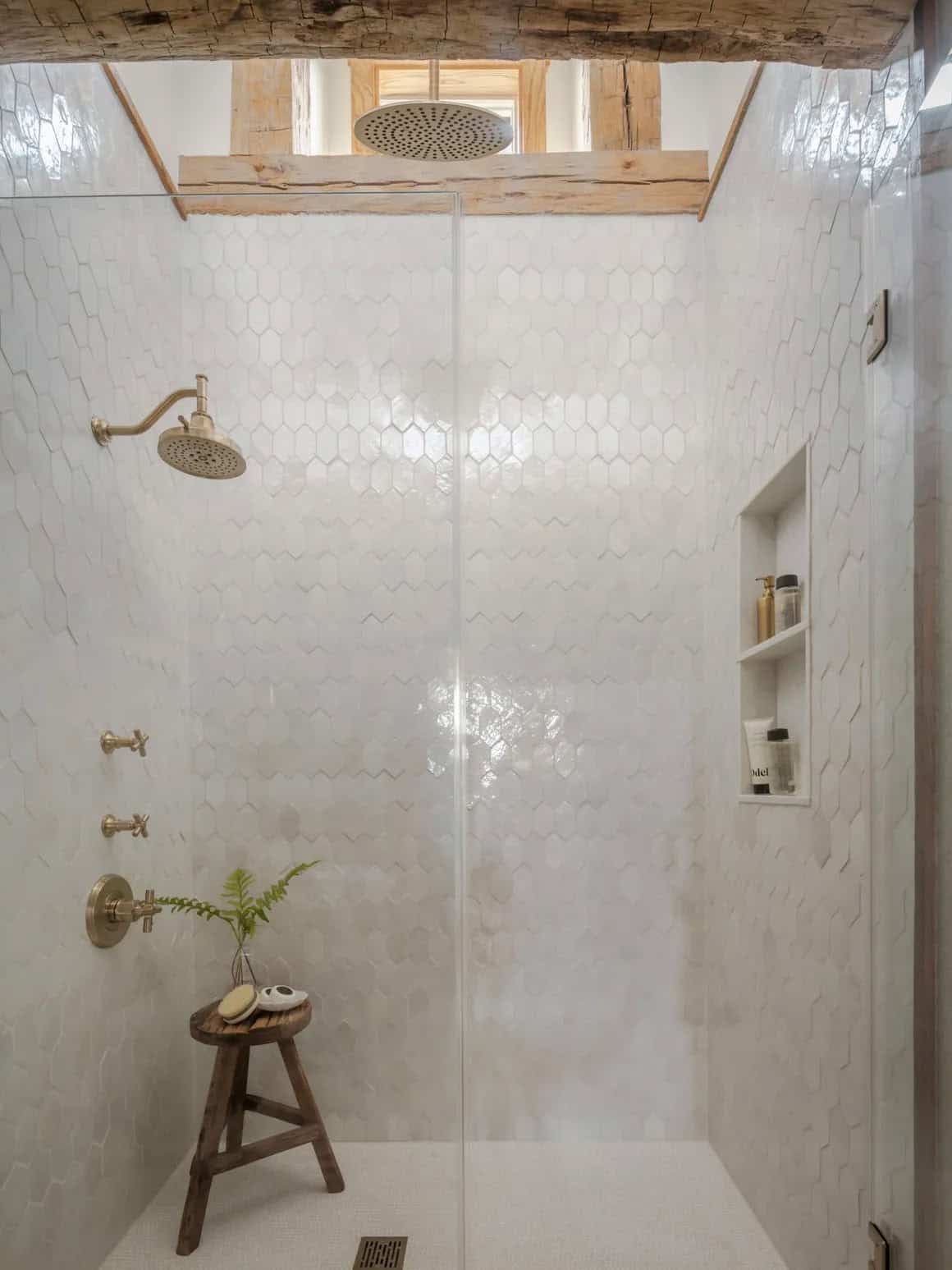 Walk-in shower with floor-to-ceiling glossy white hexagon tile walls, brass cross-handle fixtures and rain head, original reclaimed timber beam framing a small grid-pane window above, built-in niche shelving, and rustic wood stool with fern sprig