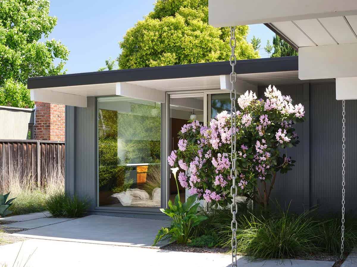 renovated Eichler home exterior with dark painted siding sliding glass doors flowering rhododendron and lush garden