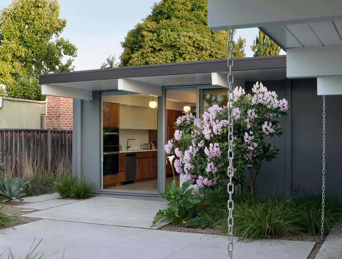 renovated Eichler home exterior at dusk with dark siding sliding glass doors to kitchen flowering rhododendron and rain chain