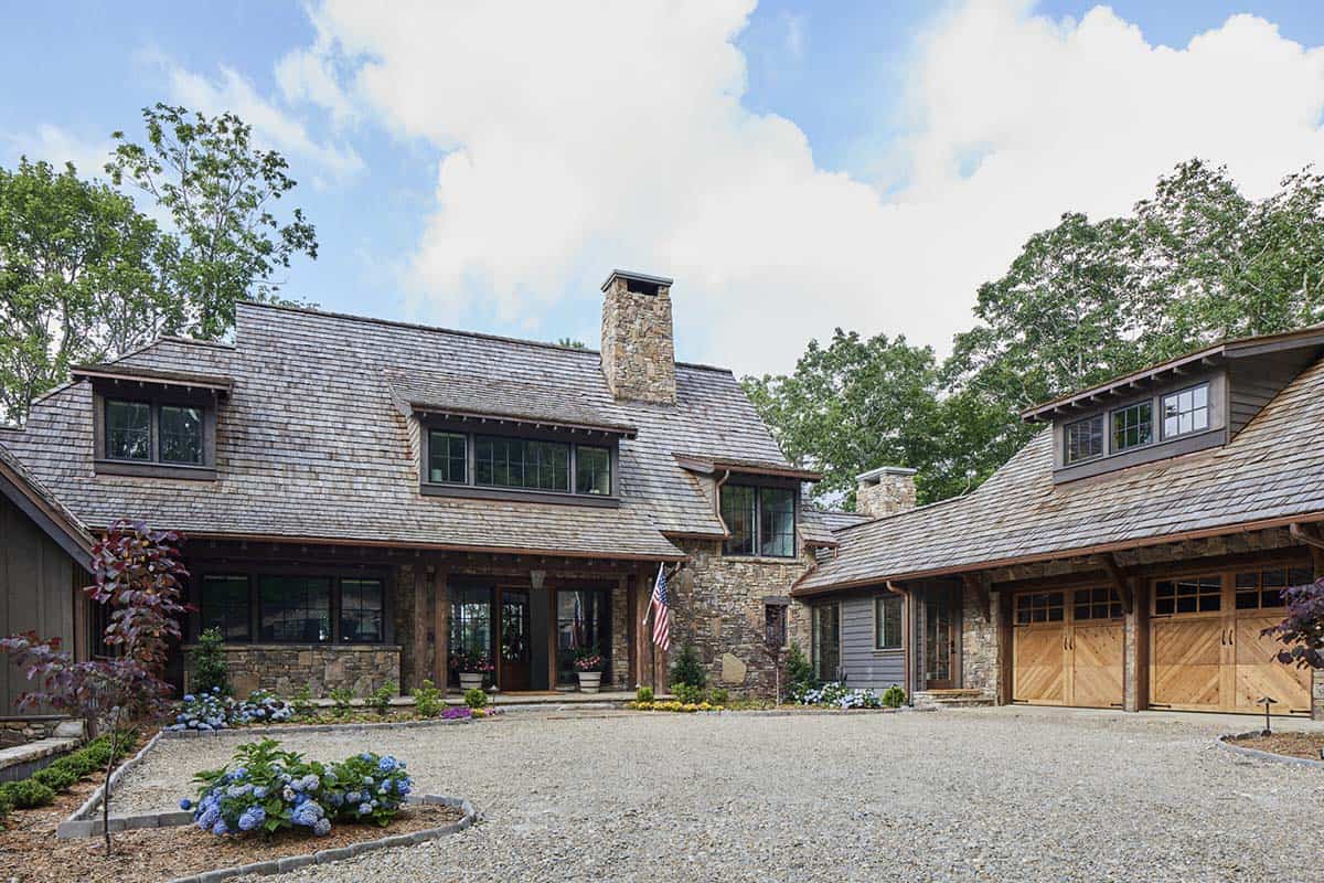 Refined mountain home exterior in North Carolina with natural fieldstone walls, cedar shake roof, warm wood entry posts, double French doors, wood garage doors, gravel motor court, and hydrangea plantings
