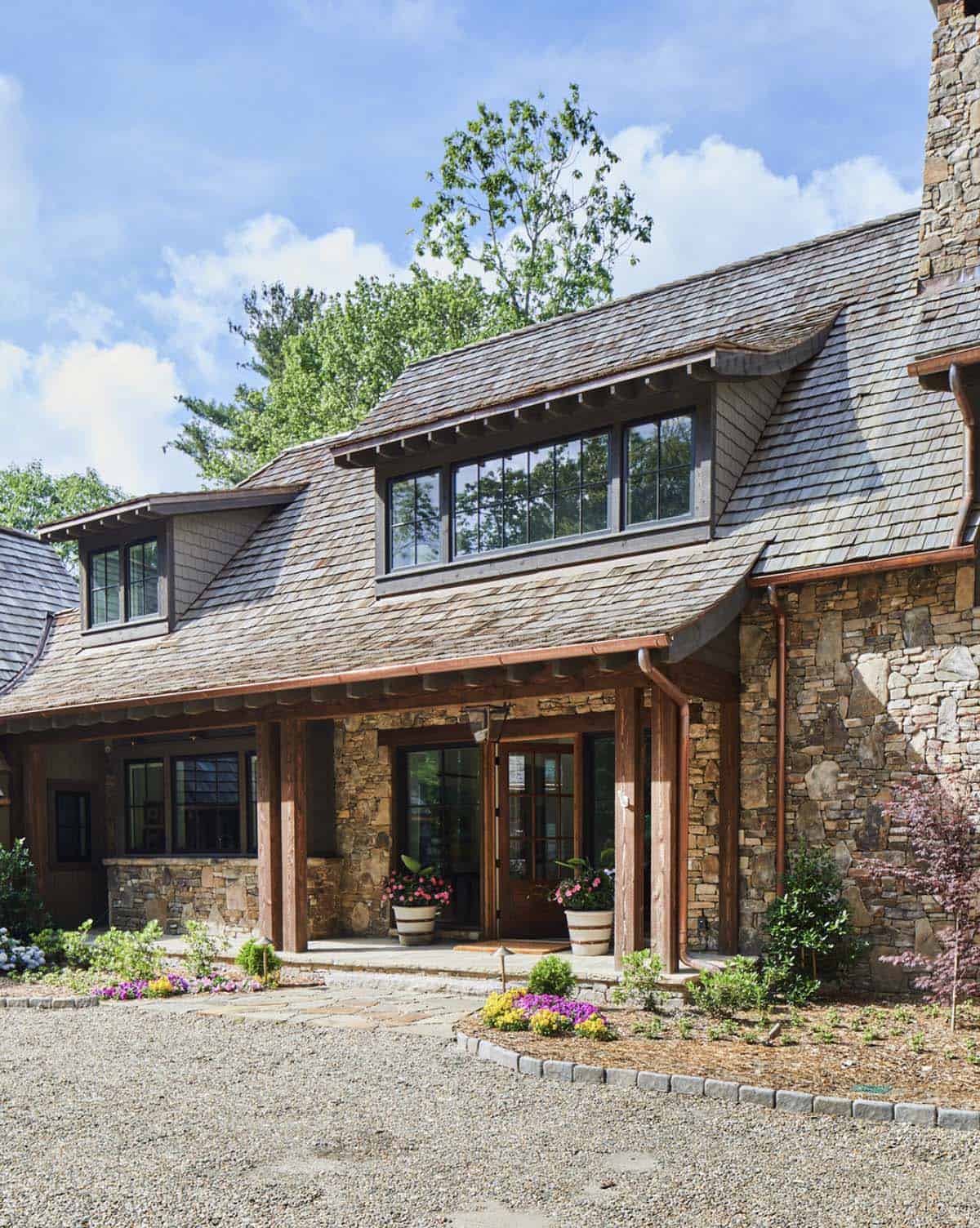 Close-up of mountain home entry with stacked stone facade, curved cedar shake roofline with eyebrow dormer, warm wood post and beam covered entry porch, French doors, and colorful flower beds