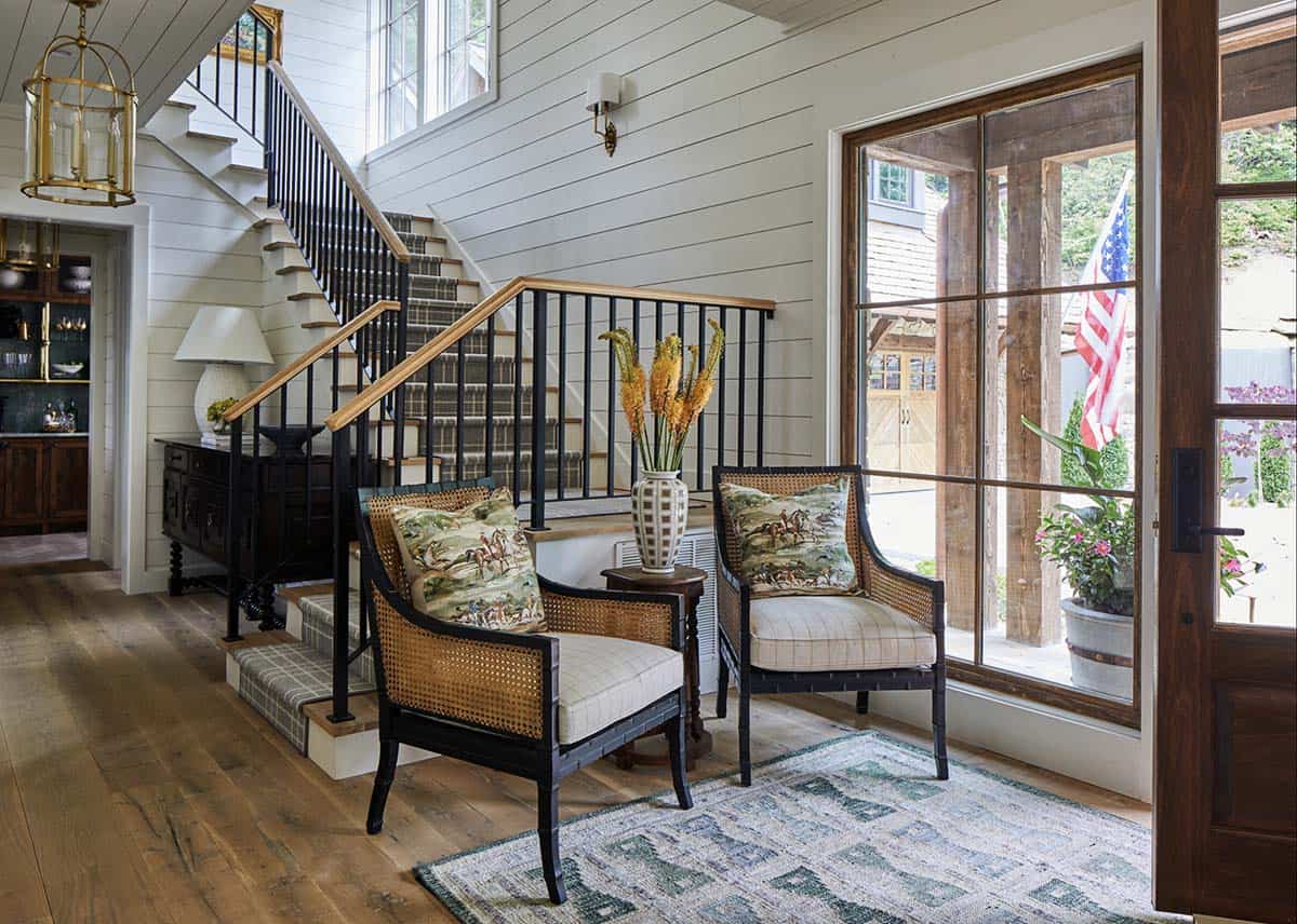 Bright entryway foyer with shiplap walls, staircase with black iron railing and plaid carpet runner, brass lantern pendant, two cane armchairs with equestrian print pillows, wide plank hardwood floors, and view to gravel courtyard through wood-framed French door