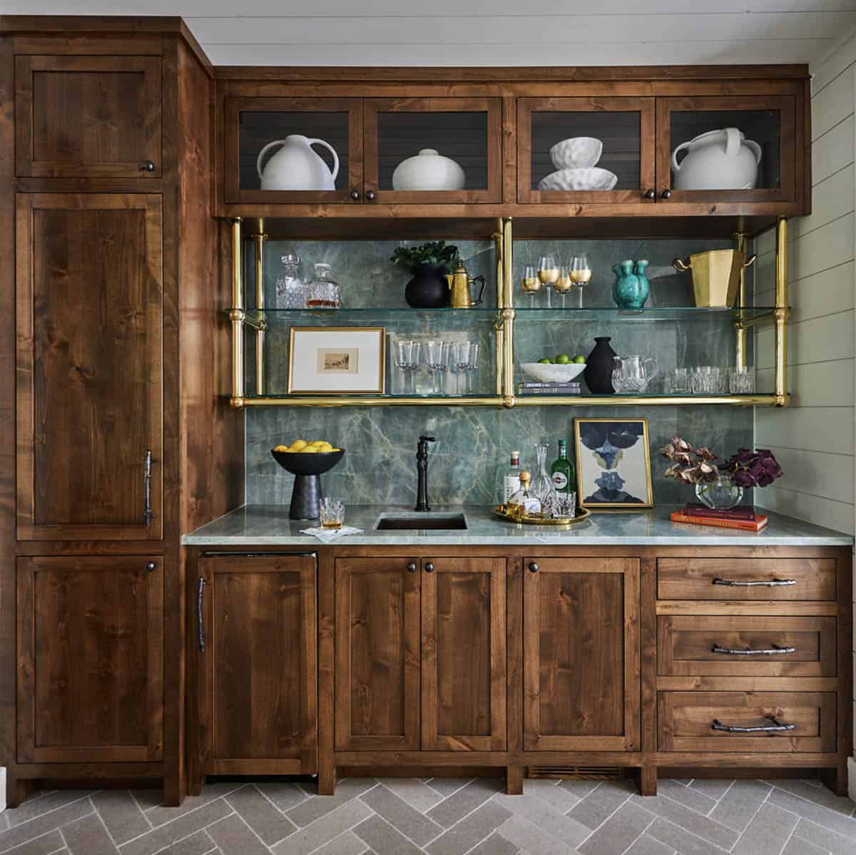 Rustic wet bar with rich walnut cabinetry, green marble slab backsplash, brass and glass open shelving, undermount bar sink with matte black faucet, marble countertop, twig-style drawer pulls, glass-front upper cabinets, and herringbone stone tile floor