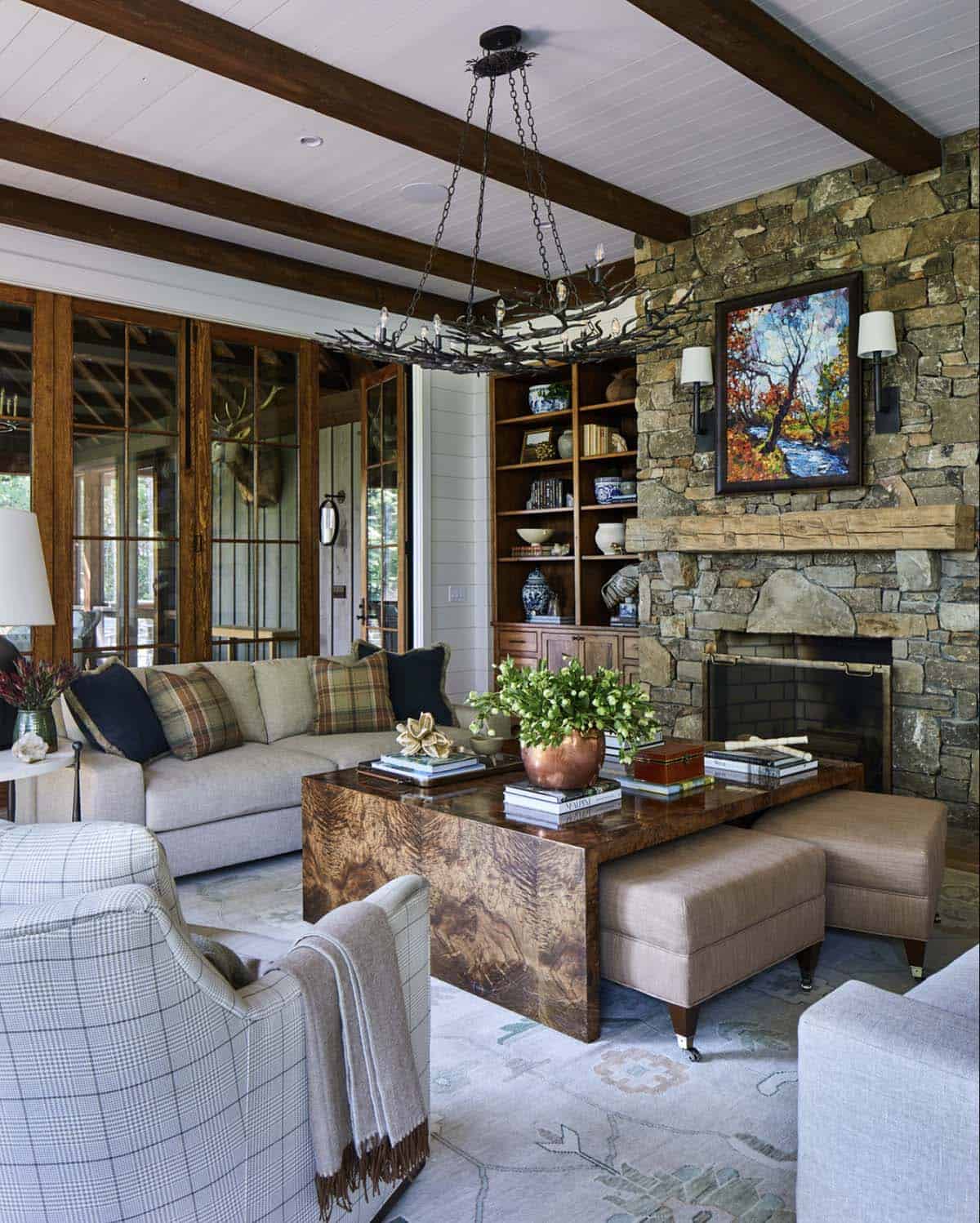 Mountain home living room with floor-to-ceiling stacked stone fireplace, reclaimed wood beam ceiling, twig and iron chandelier, burl wood coffee table, gray upholstered seating with plaid pillows, built-in wood bookcase, and wood and glass French doors to screened porch