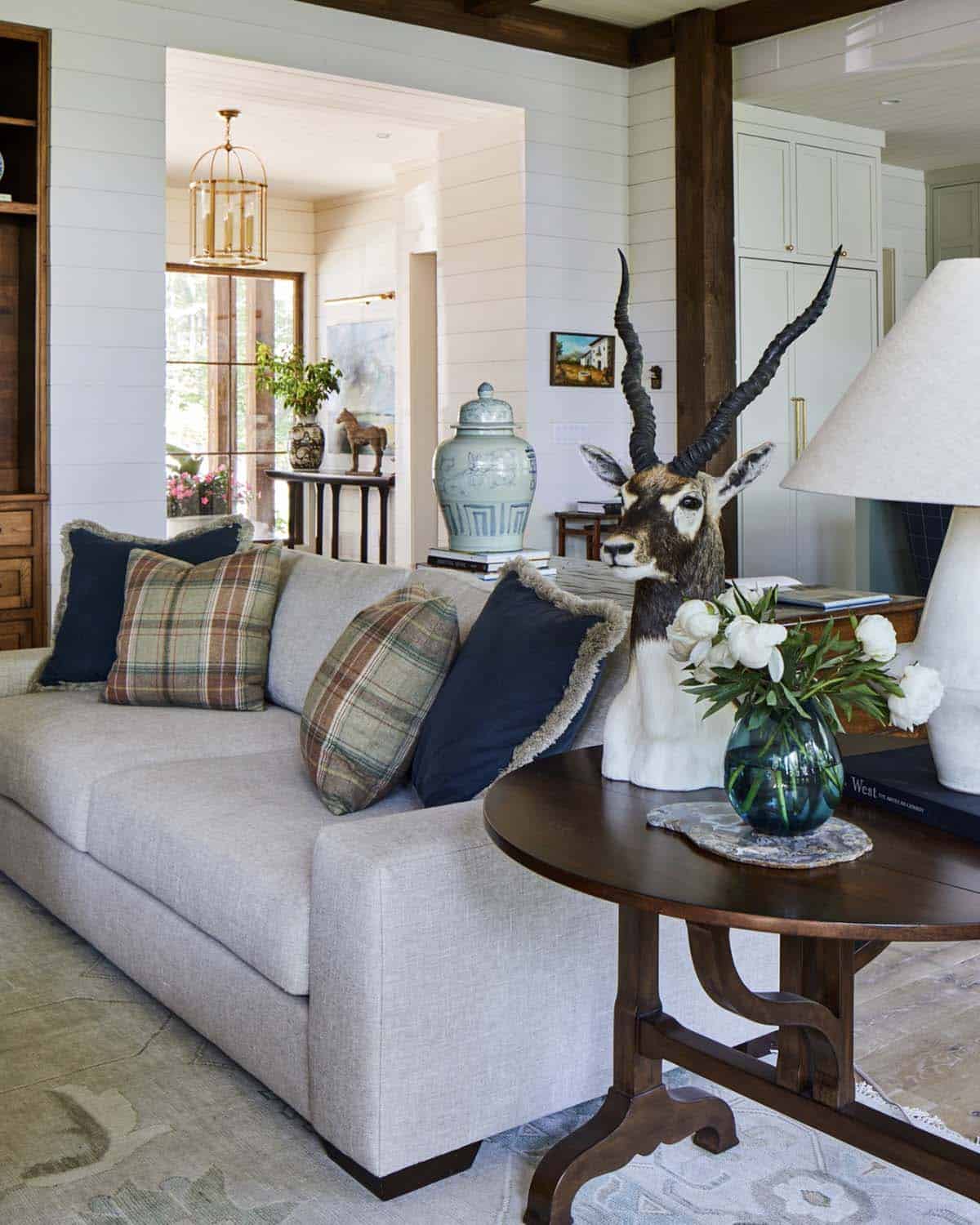 Living room detail with gray linen sofa, plaid and navy fringe accent pillows, round dark wood side table, antelope mount, celadon ginger jar, white peony arrangement in blue glass vase, and view through doorway to entryway foyer