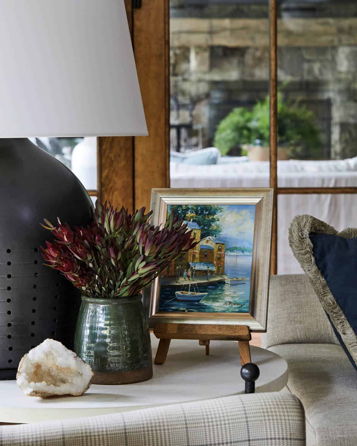 Living room side table vignette with green glazed ceramic vase of burgundy protea stems, small framed harbor oil painting on miniature easel, raw crystal geode, black perforated lamp base, and reflection of stone and wood windows