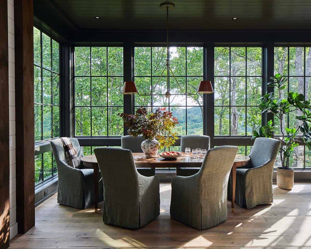 Dramatic mountain home dining room wrapped in floor-to-ceiling black-framed grid windows overlooking lush North Carolina forest, round wood dining table, gray slipcovered chairs, dual red shade pendant light, Japanese maple branch centerpiece in chinoiserie vase, and fiddle leaf fig tree
