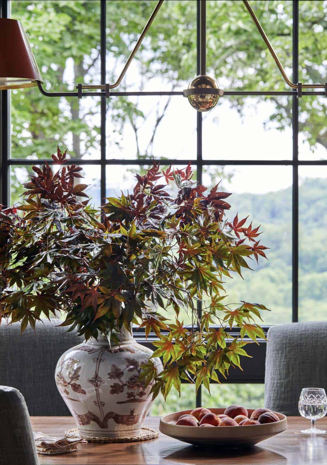 Close-up of dining table centerpiece with Japanese maple branches in white chinoiserie vase, wooden bowl of fresh peaches, crystal wine glass, linen napkin, and brass and black dual pendant light against floor-to-ceiling black grid windows with mountain forest views
