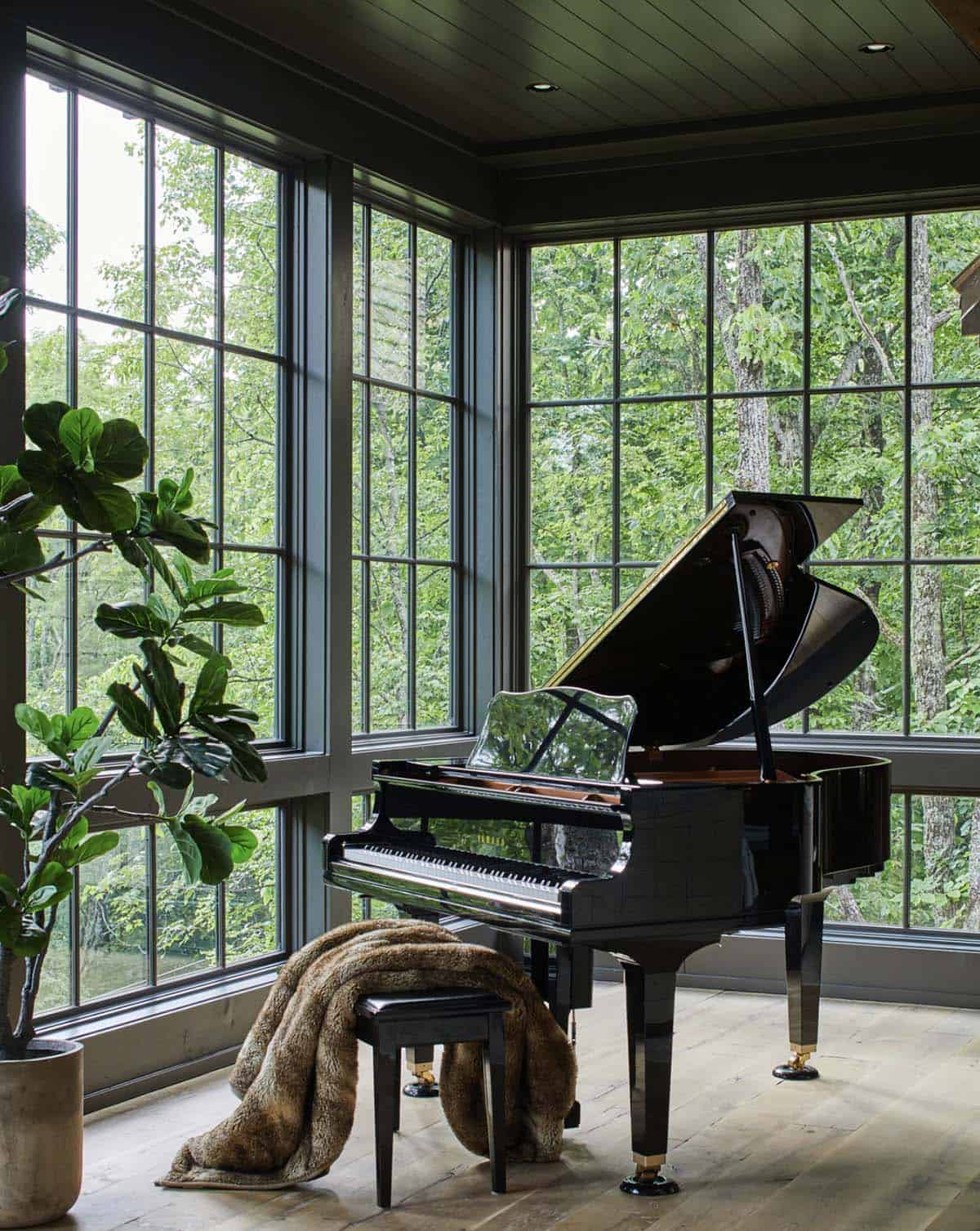 Grand piano room with floor-to-ceiling black-framed grid windows on two walls overlooking dense green North Carolina woodland, black lacquer baby grand piano with fur throw on bench, fiddle leaf fig tree, and wide plank hardwood floors