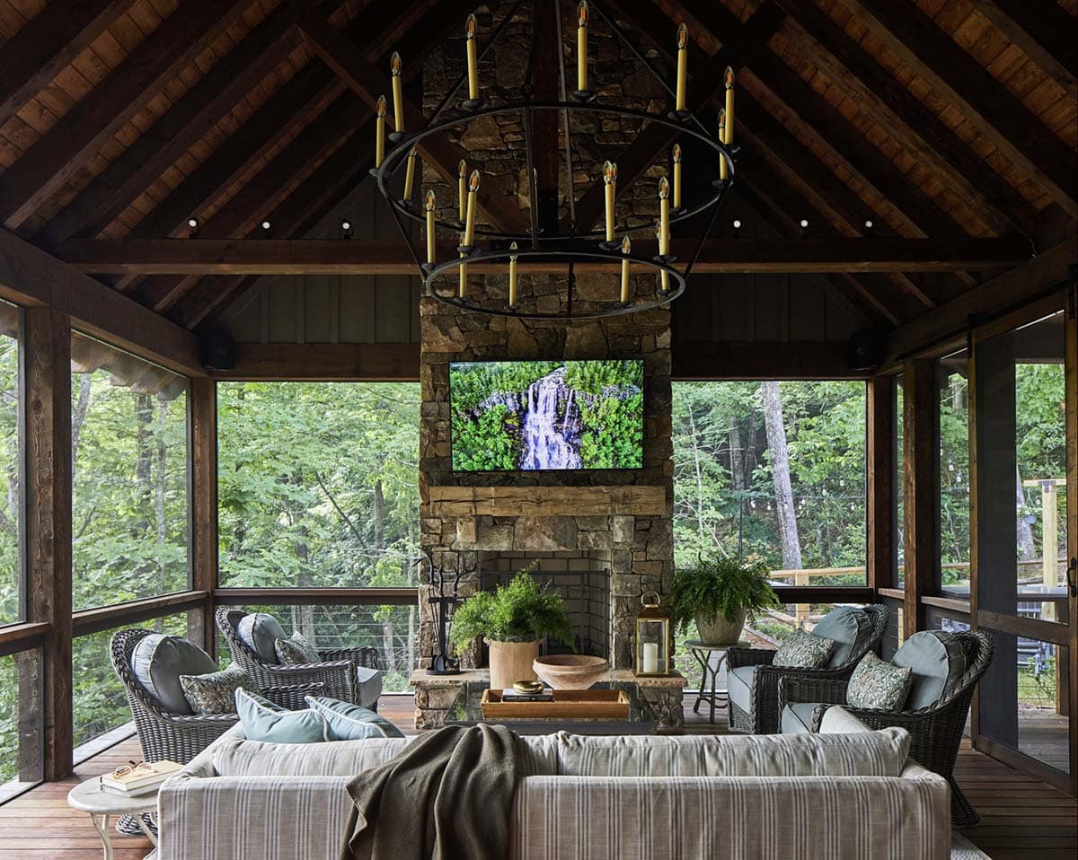 Covered screened porch with vaulted dark wood beam ceiling, large iron ring candle chandelier, stacked stone fireplace with mounted outdoor television, wicker and upholstered seating in blue and gray, and panoramic views of surrounding North Carolina forest
