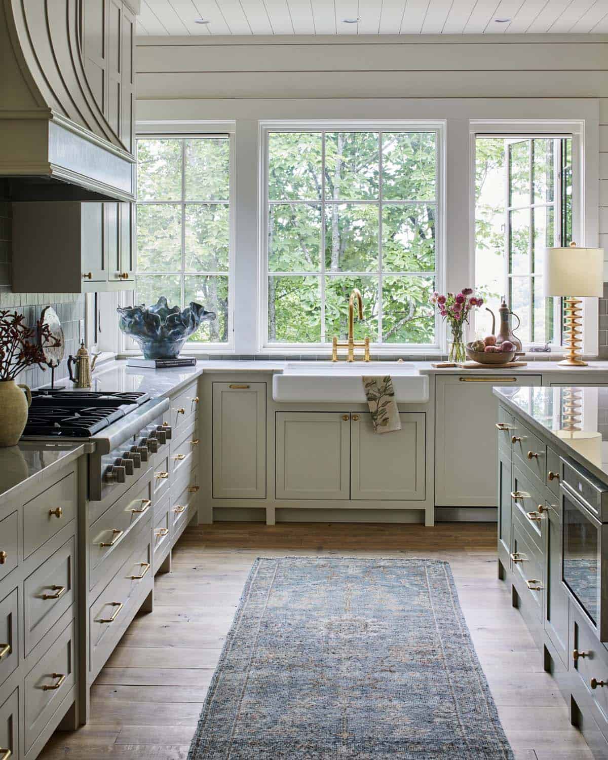 Mountain home kitchen with sage green shaker cabinetry, brass hardware, white farmhouse apron sink, marble countertops, sage green curved range hood, grid-pane windows overlooking treetops, wide plank hardwood floors, and vintage blue runner rug