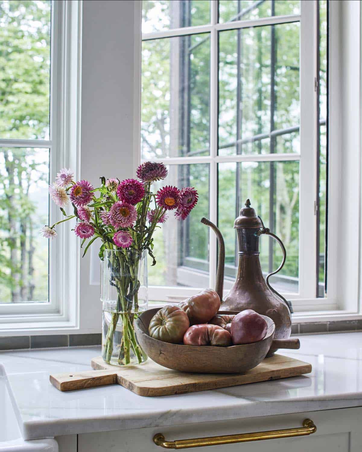 Kitchen counter vignette with pink strawflowers in glass vase, heirloom tomatoes in wooden dough bowl on cutting board, antique copper ewer, marble countertop with brass pull, and open grid-pane window with lush green mountain views
