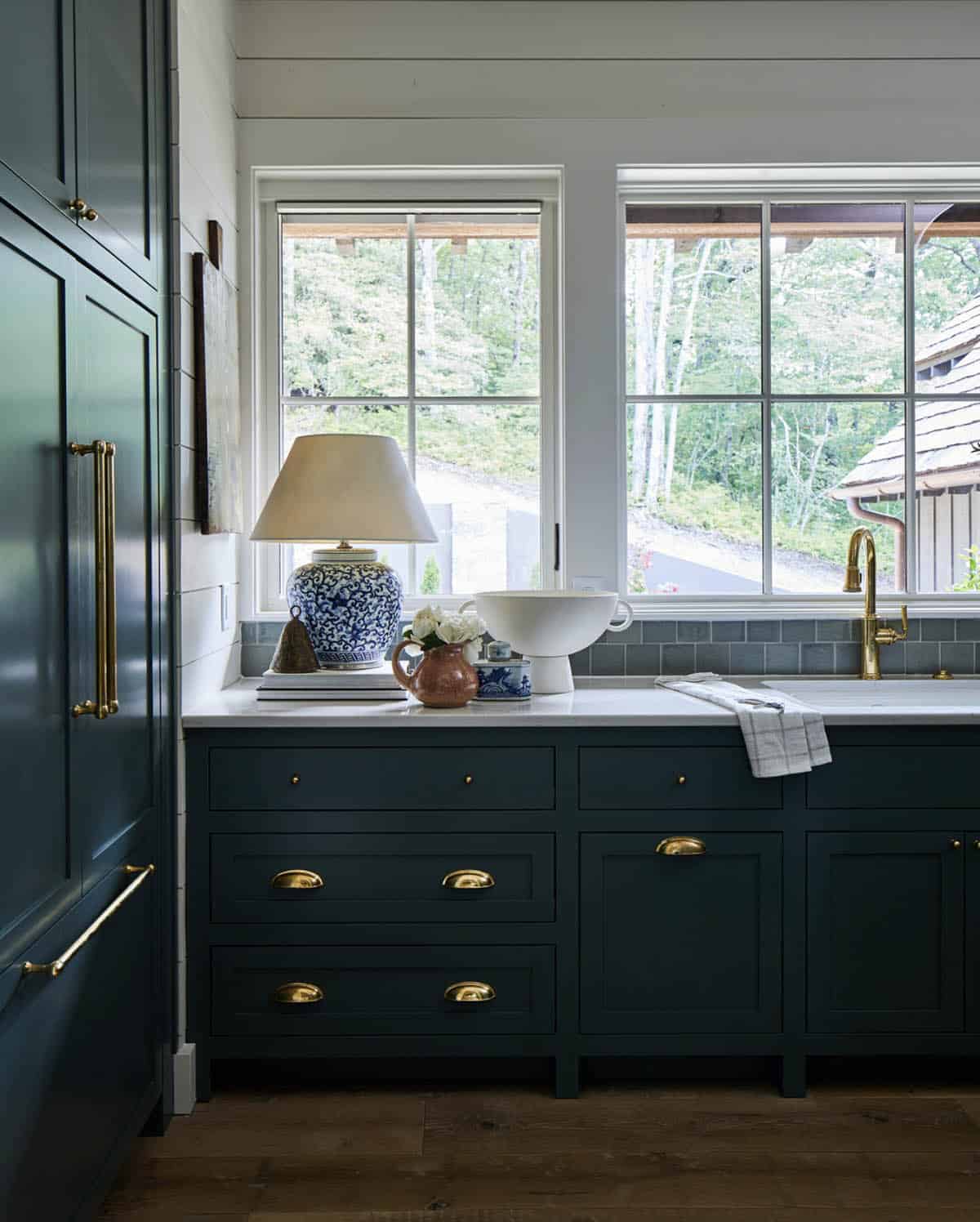 Dark teal kitchen cabinets with brass cup pulls and bar handles, white marble countertop, gray subway tile backsplash, brass faucet, and blue-and-white ginger jar lamp beside sink window overlooking forest