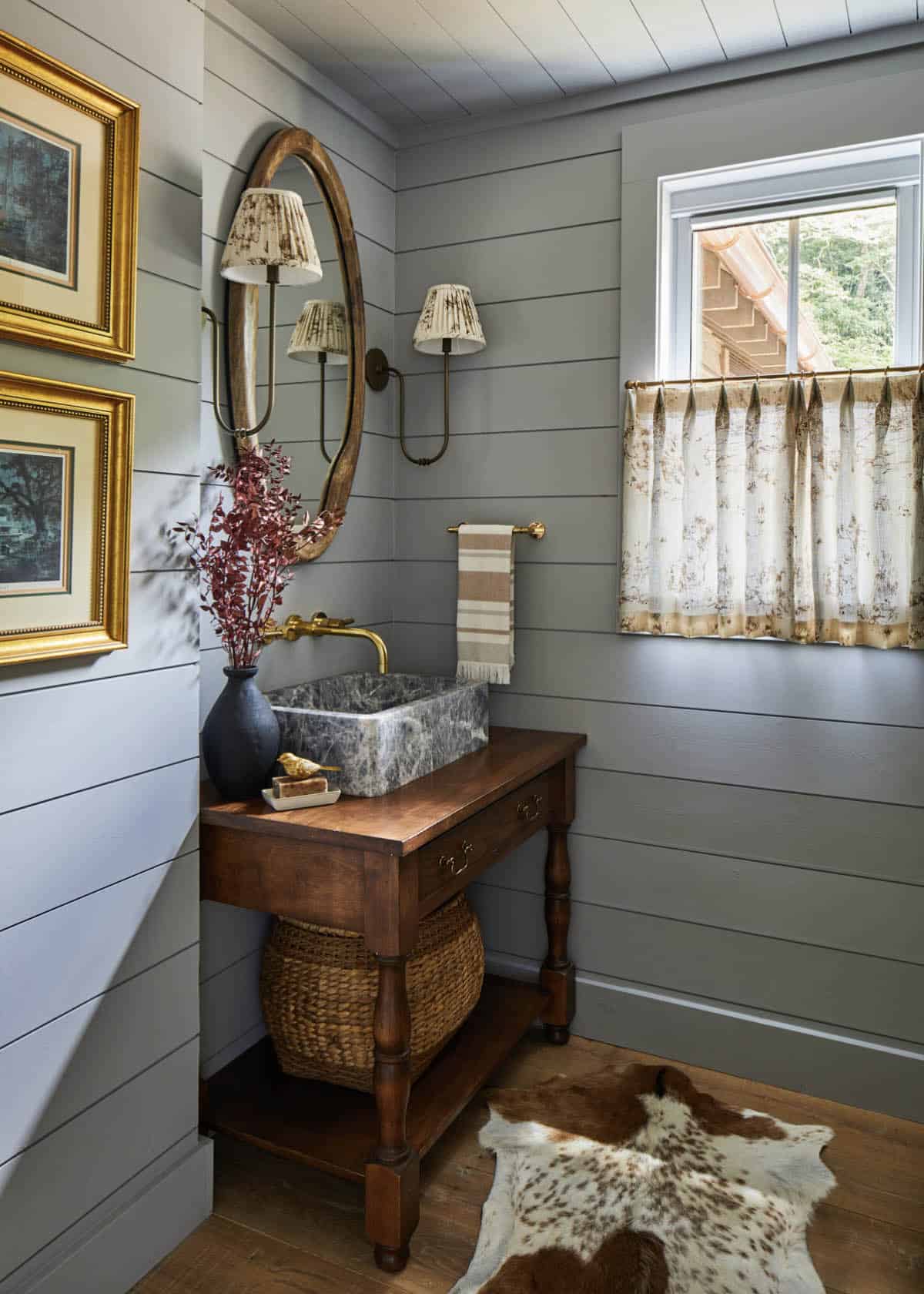 Powder room with gray shiplap walls, antique wood vanity, honed marble vessel sink, brass wall-mount faucet, oval gilt mirror, brass sconce with pleated shades, cowhide rug, and floral caf&eacute; curtains