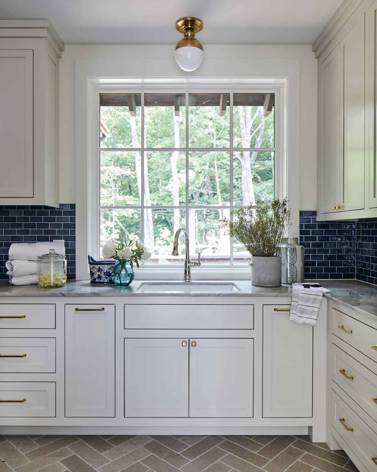 Laundry room with cream shaker cabinets, navy blue subway tile backsplash, gray marble countertop, brass hardware, herringbone slate floor tile, and a large window overlooking trees