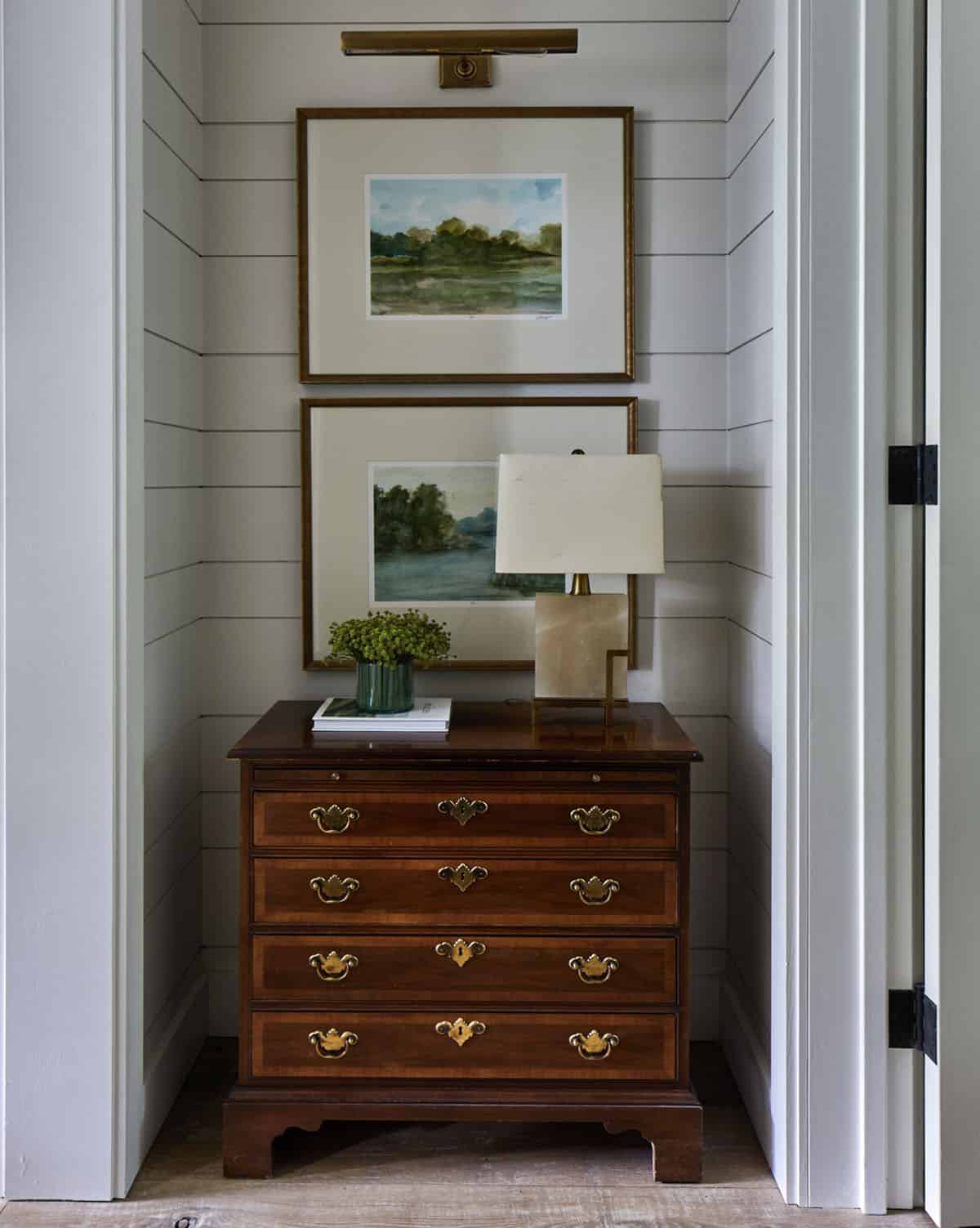 Hallway niche with gray shiplap walls, antique mahogany four-drawer chest with brass bail pulls, brass picture light, two watercolor landscape paintings in gilt frames, and alabaster table lamp