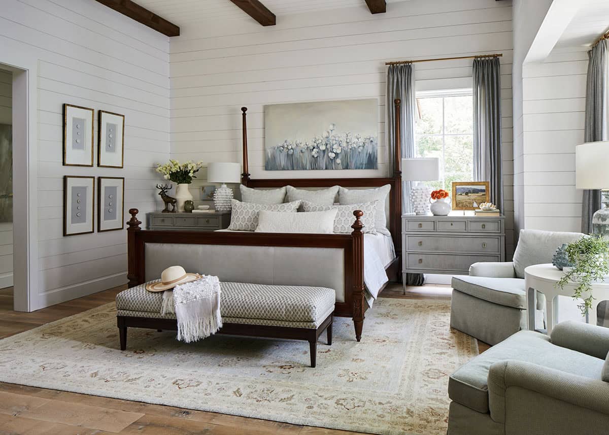 Serene bedroom with white shiplap walls, dark wood exposed ceiling beams, mahogany four-poster bed with upholstered linen panel, gray sage armchairs, vintage oushak rug, and floral abstract canvas