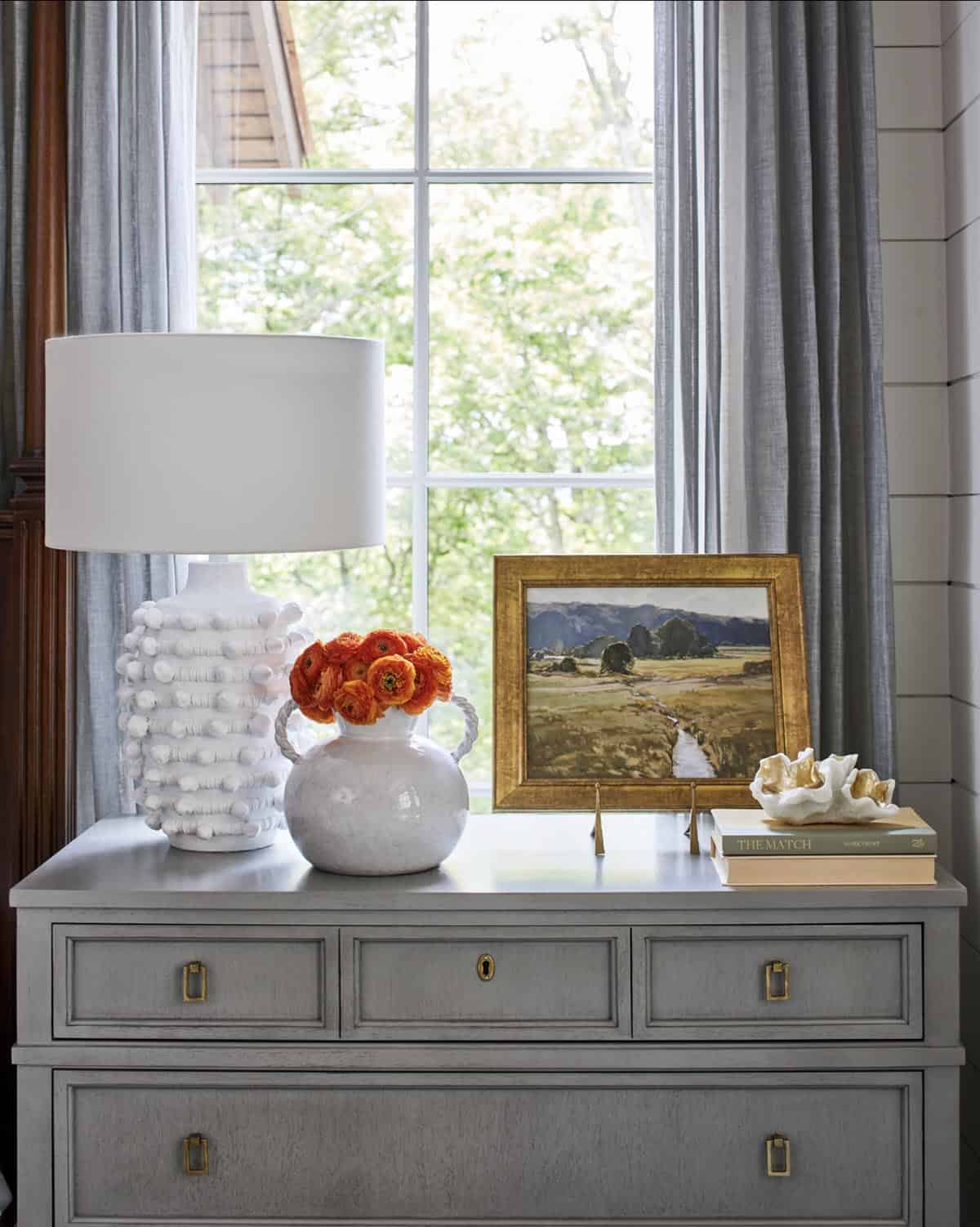 Bedroom nightstand vignette with gray painted dresser, white ceramic bobble lamp, round white ceramic vase with orange ranunculus, small landscape oil painting in gilt frame, and gray linen curtains