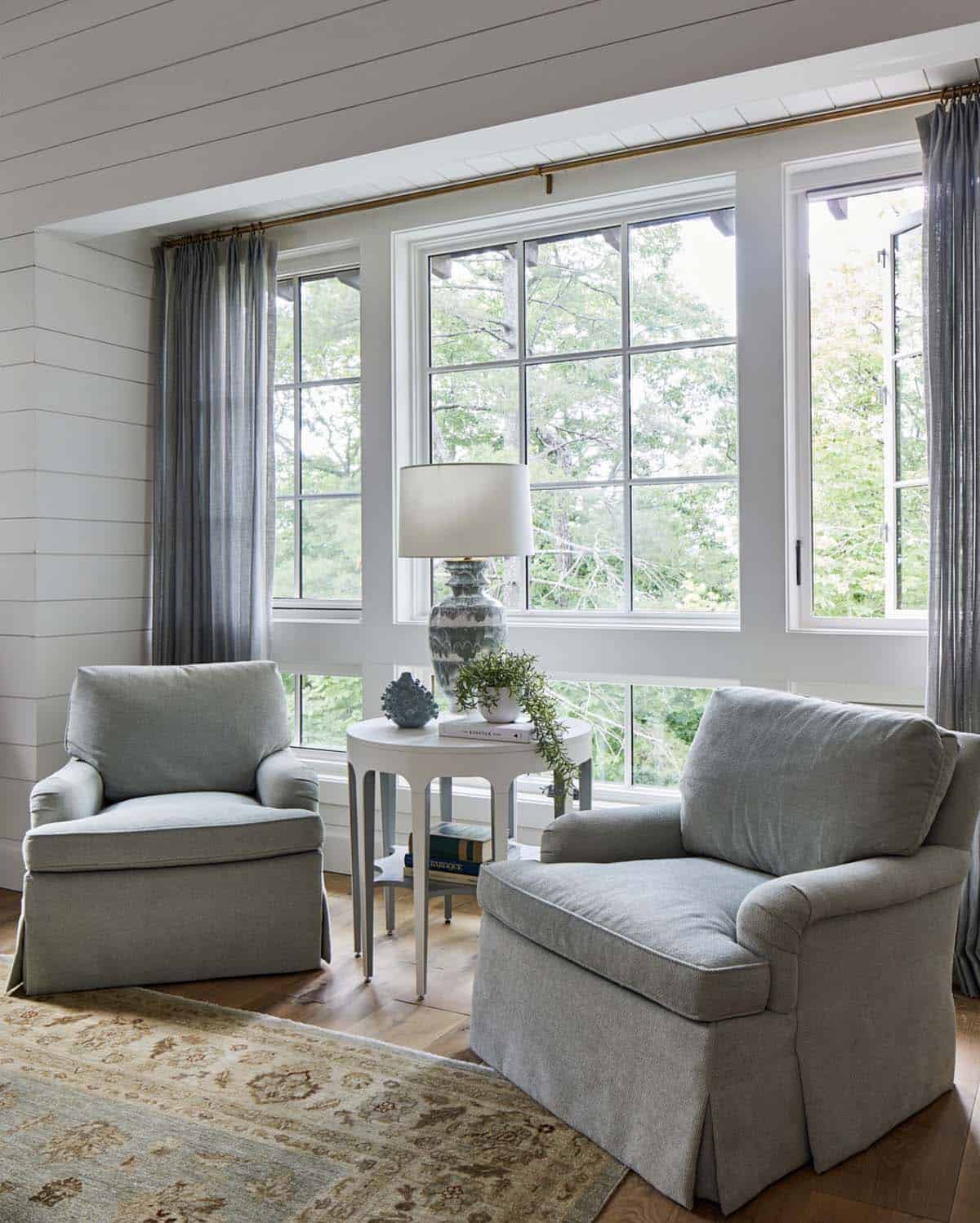 Bedroom sitting area with two sage green upholstered swivel chairs, white side table, blue-and-white ceramic lamp, gray linen curtains on brass rod, shiplap walls, and floor-to-ceiling wooded view