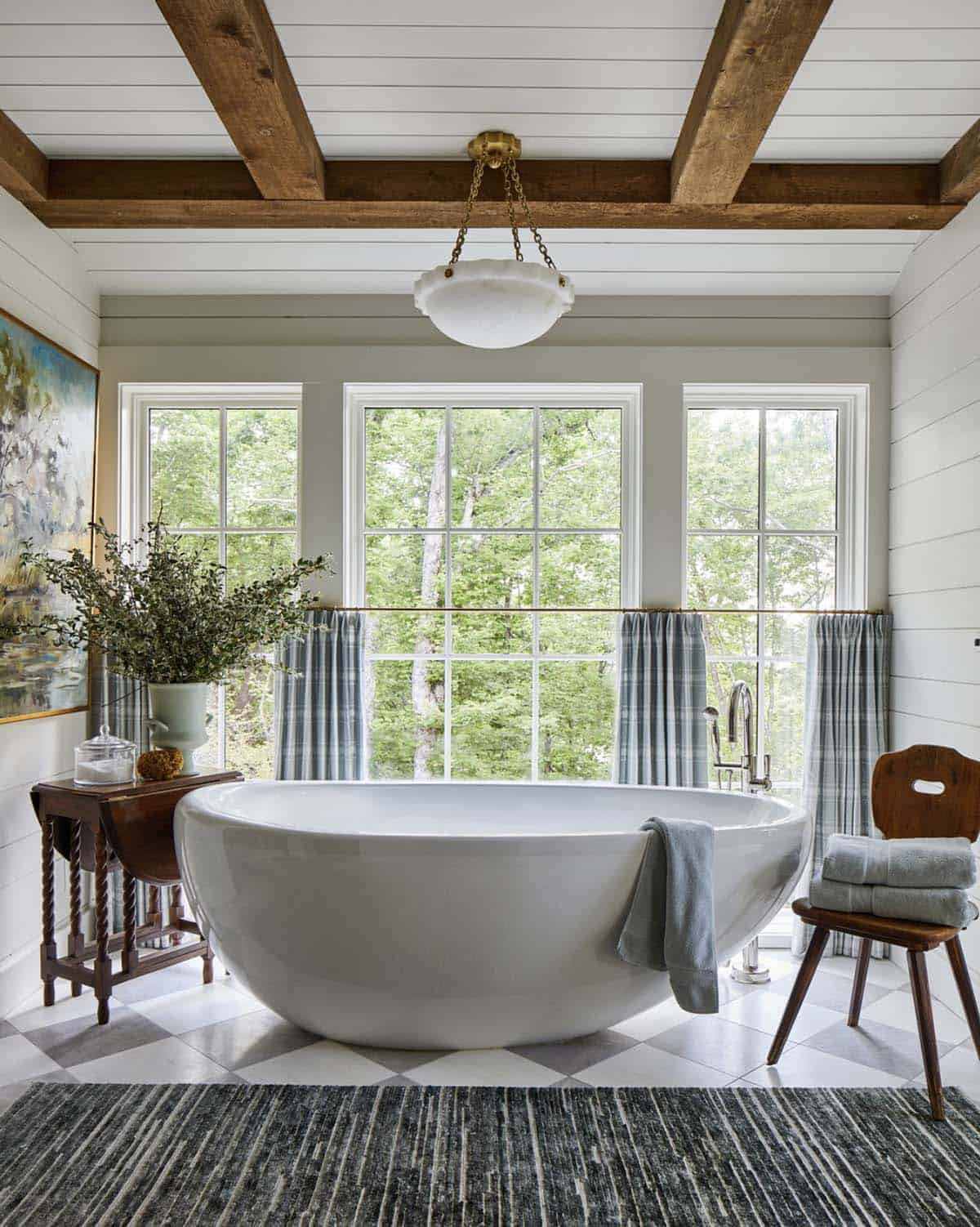 Spa-like bathroom with oval freestanding soaking tub, gray-and-white harlequin tile floor, white shiplap walls, exposed wood ceiling beams, three large windows with plaid caf&eacute; curtains, and brass chain pendant light