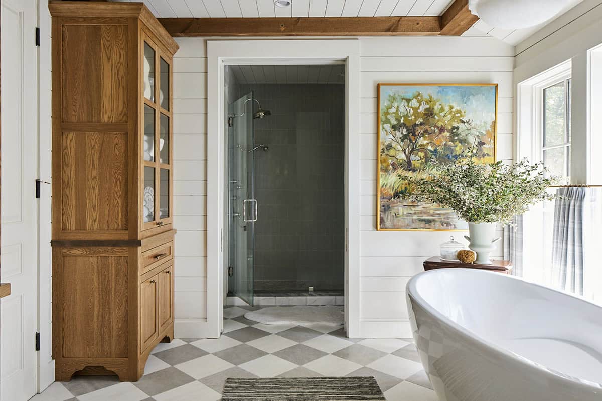 Bathroom with harlequin tile floor, freestanding soaking tub, walk-in shower with dark green tile, antique oak linen armoire with glass doors, white shiplap walls, and impressionist tree painting