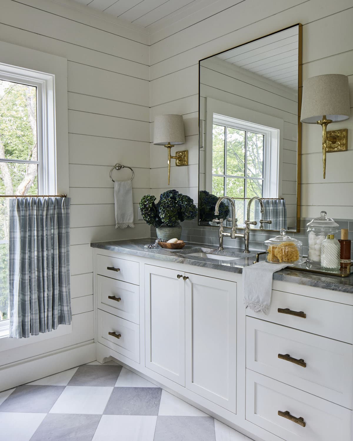 Bathroom vanity with white shaker cabinets, soapstone countertop, polished nickel bridge faucet, brass-framed tall mirror, linen sconces on brass brackets, plaid caf&eacute; curtains, and harlequin tile floor