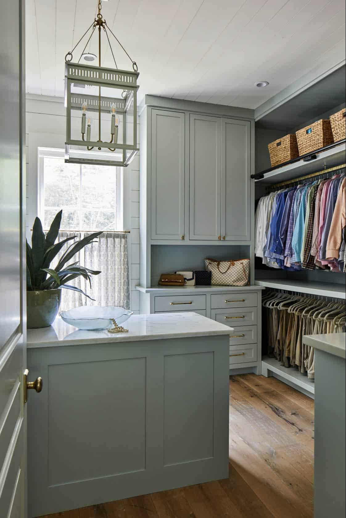 Walk-in closet with sage green built-in cabinetry, white marble island, brass lantern chandelier, open hanging rods with organized clothing, woven storage baskets, and shiplap walls with caf&eacute; curtains