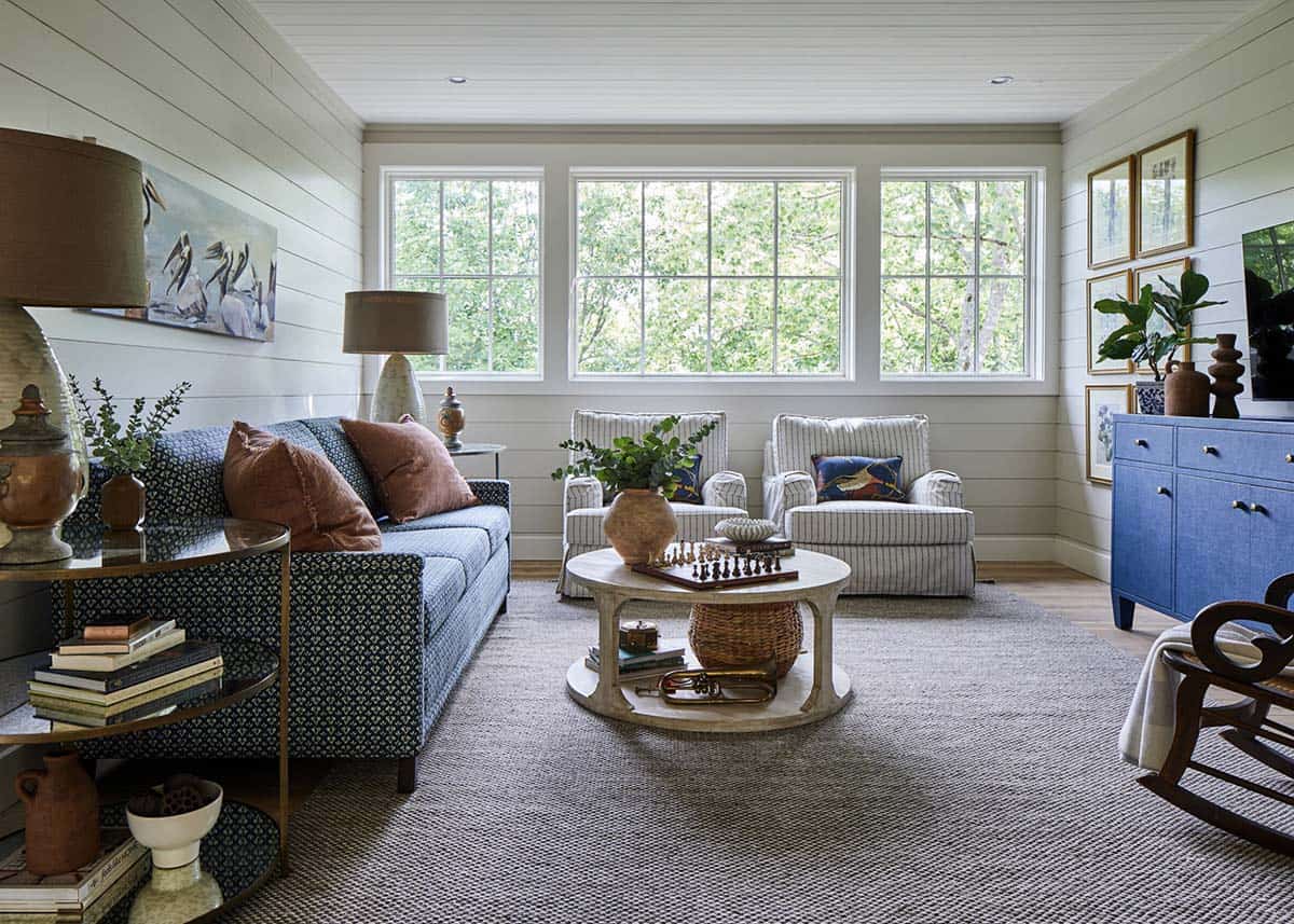 Casual family room with blue printed sofa, rust velvet pillows, striped slipcovered chairs, round whitewashed coffee table, blue grasscloth credenza, shiplap walls, triple picture windows, and gallery wall