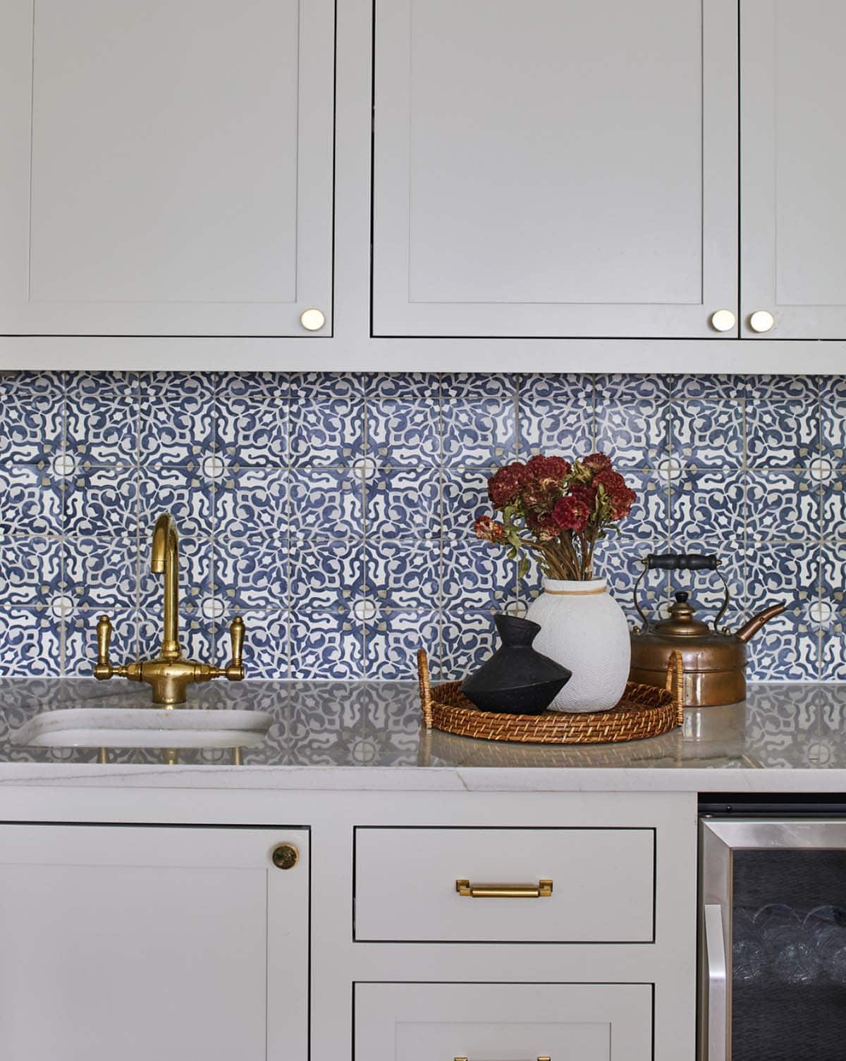Wet bar with light gray shaker cabinets, brass hardware, bold blue-and-white Moroccan encaustic tile backsplash, marble countertop, brass bridge faucet, wicker tray vignette, and wine refrigerator