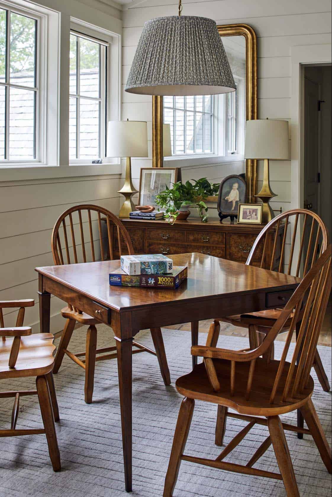 Game room with antique mahogany drop-leaf table, Windsor chairs, woven pendant shade, burled wood sideboard styled with vintage brass lamps, gold mirror, family photos, and shiplap walls
