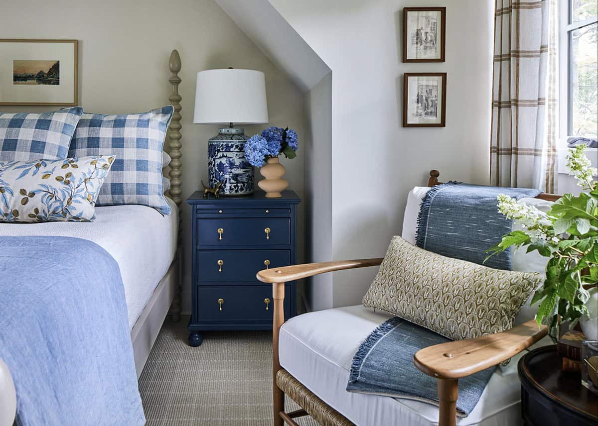 Guest bedroom with spindle bed, blue buffalo check and floral pillows, navy painted nightstand with brass pulls, blue-and-white ginger jar lamp, plaid curtains, and vaulted ceiling with angled wall
