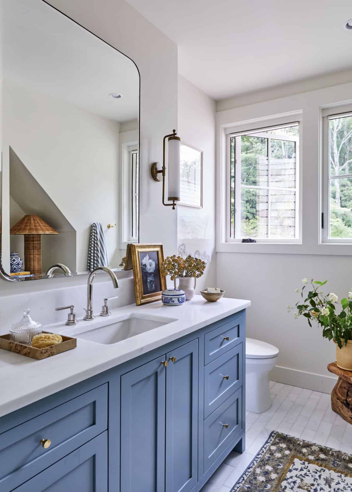 Guest bathroom with slate blue shaker vanity, white quartz countertop, nickel widespread faucet, large frameless mirror, brass tube sconce, vintage rug, and windows with wooded view