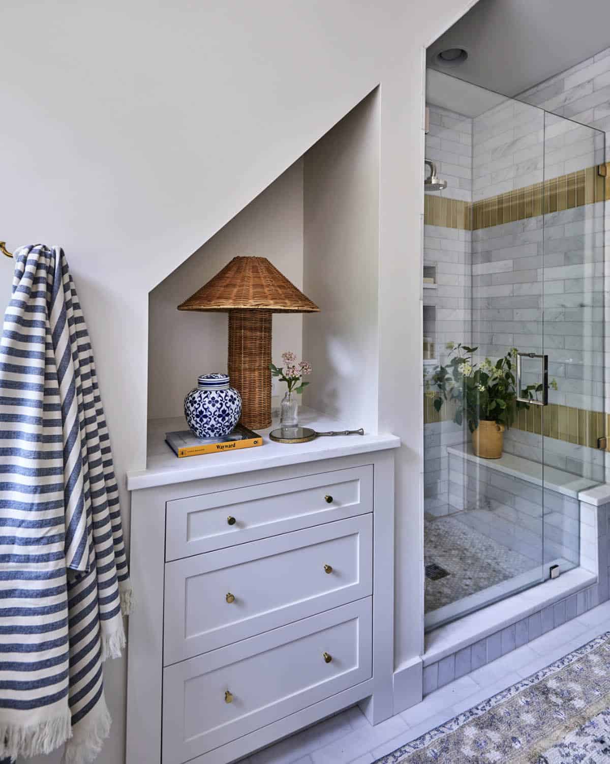 Bathroom with built-in storage under angled roofline, white shaker drawers with brass knobs, wicker lamp, blue-and-white ginger jar, walk-in glass shower with marble and olive green vertical tile stripe detail