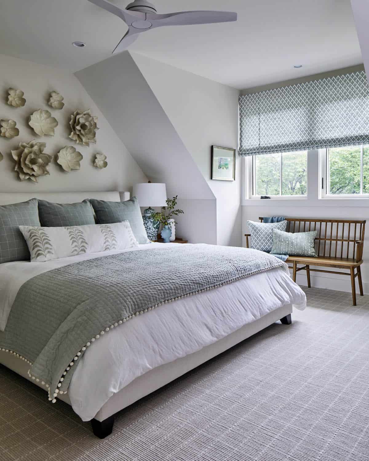 Guest bedroom with vaulted ceiling, white upholstered platform bed, sage green quilted coverlet with pom-pom trim, ceramic flower wall art, walnut bench, patterned roman shades, and ceiling fan