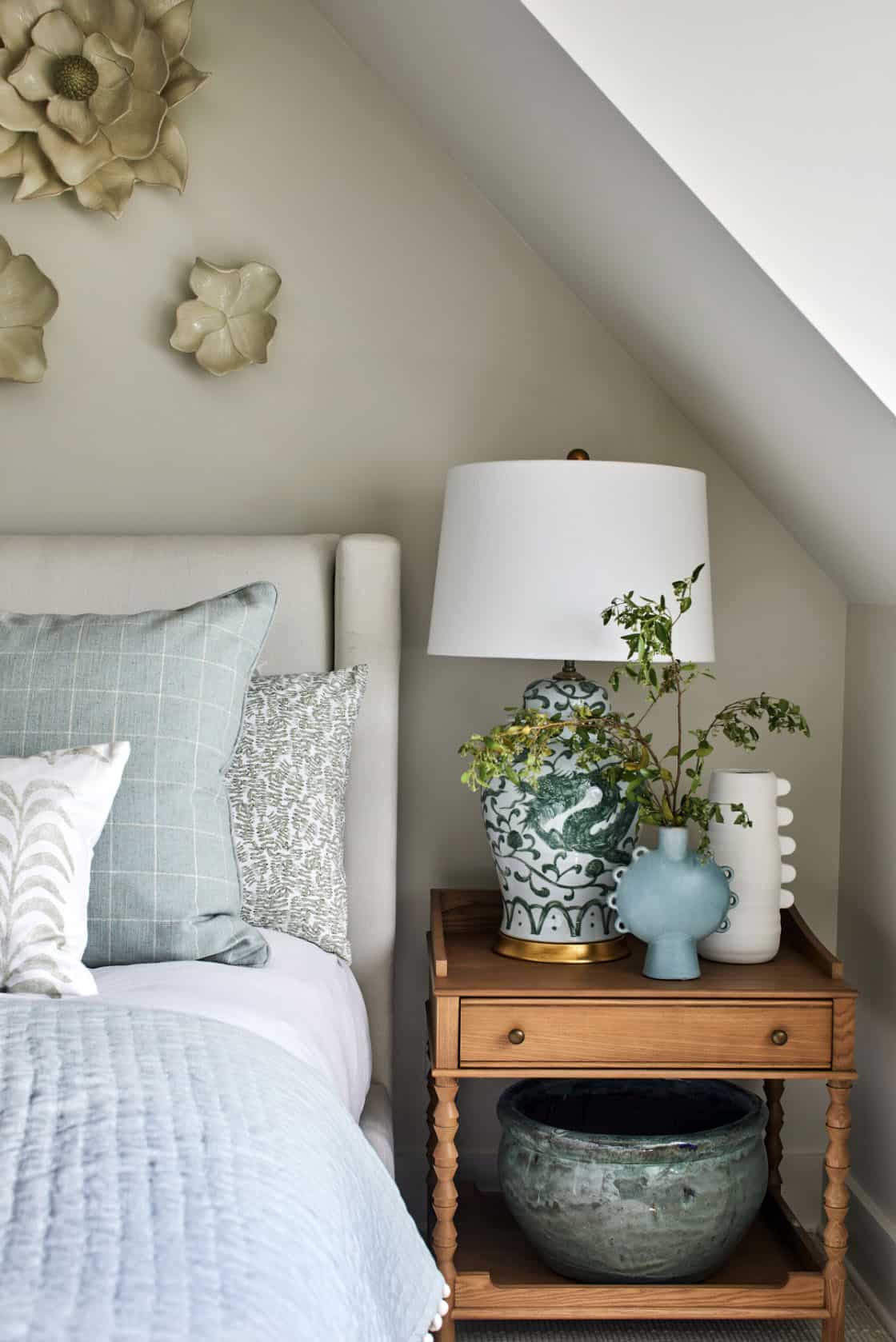 Bedroom nightstand vignette with walnut bobbin side table, green-and-white dragon ginger jar lamp, blue ceramic bud vase, white ribbed vase, textured throw pillows, and ceramic flower wall medallions above