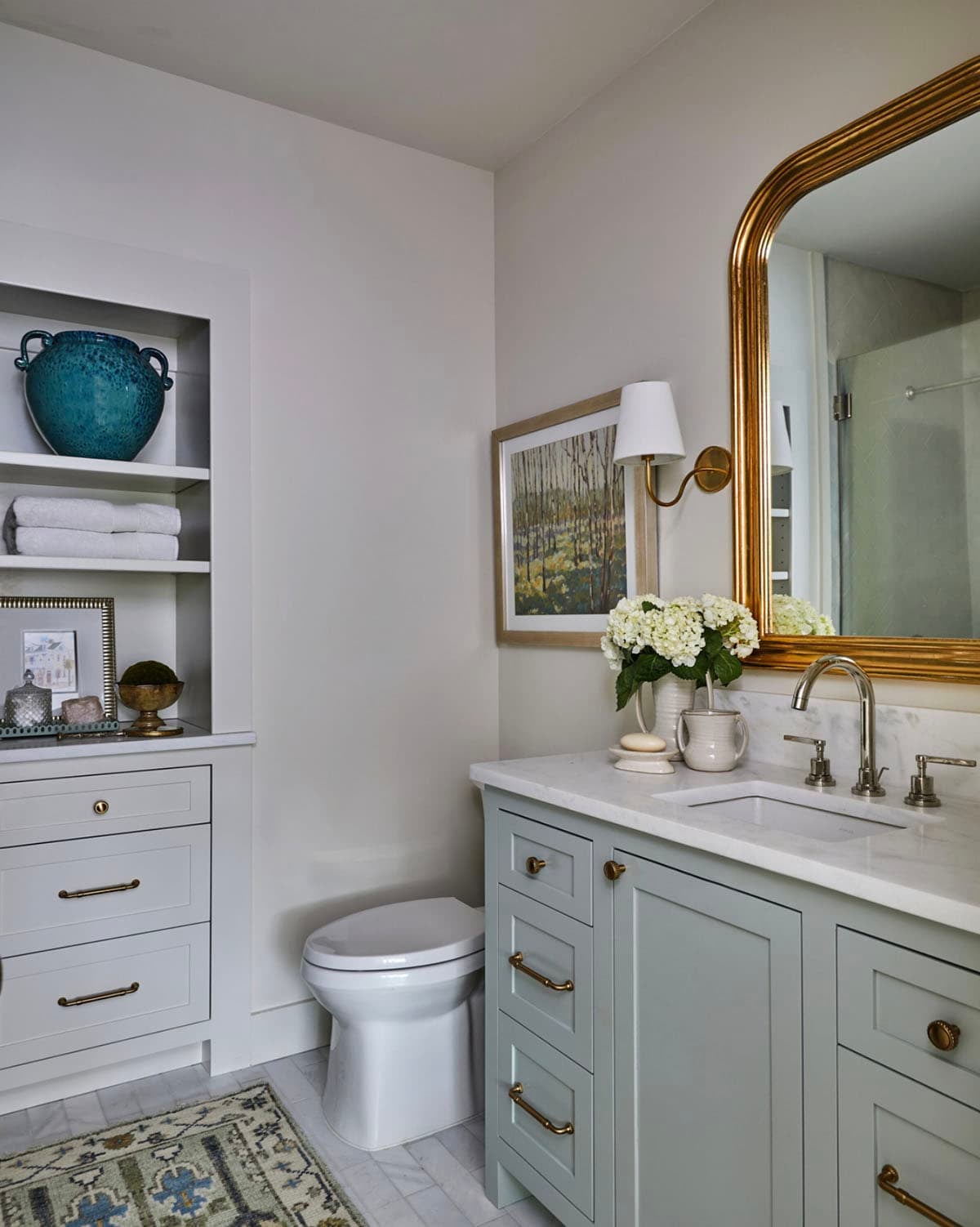 Bathroom with sage green shaker vanity, marble countertop, nickel widespread faucet, ornate gold mirror, brass swing-arm sconce, built-in niche shelving with blue ceramic vessel, and vintage rug