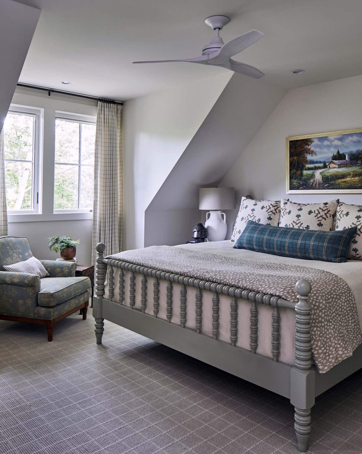 Guest bedroom with vaulted ceiling, gray painted spindle bed, antelope print bedding, plaid bolster pillow, floral upholstered chair, plaid curtains, ceiling fan, and pastoral oil painting above headboard