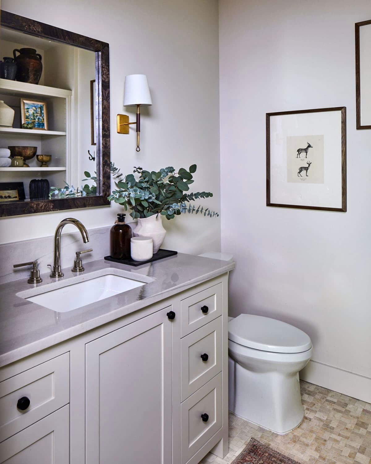 Small guest bathroom with white shaker vanity, gray quartz countertop, black ceramic knobs, brushed nickel faucet, tortoiseshell mirror, brass swing-arm sconce, eucalyptus stems, antique botanical deer prints, and mosaic tile floor