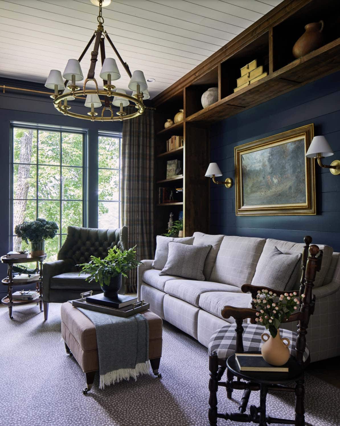 Sophisticated navy study with brass ring chandelier, plaid upholstered sofa, tufted green leather wingback chair, plaid armchair, rustic wood built-in bookshelves, equestrian oil painting in gilt frame, and plaid curtains